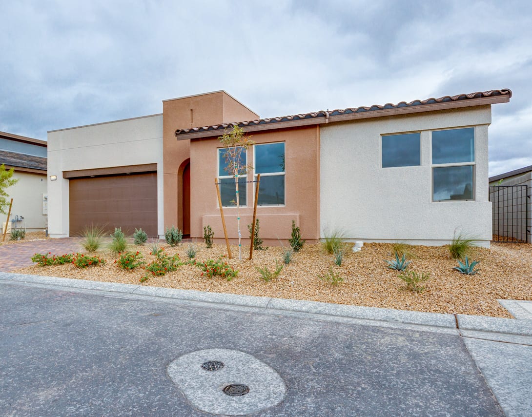 A modern, single-story house with a tiled roof, surrounded by a gravel driveway and desert landscaping, set against a cloudy sky.