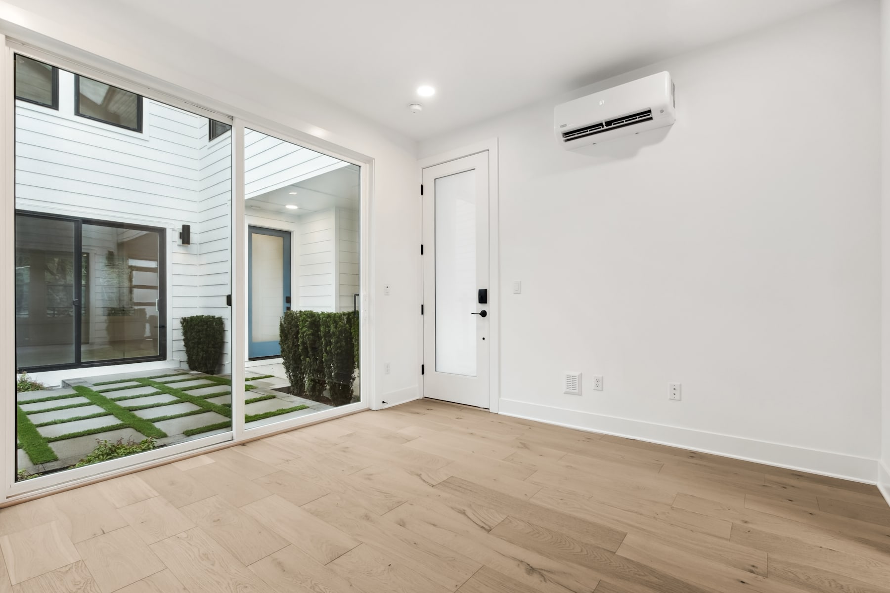 A bright, modern interior with a sliding glass door leading to a small outdoor area with greenery, and a white wall with a mounted air conditioning unit.