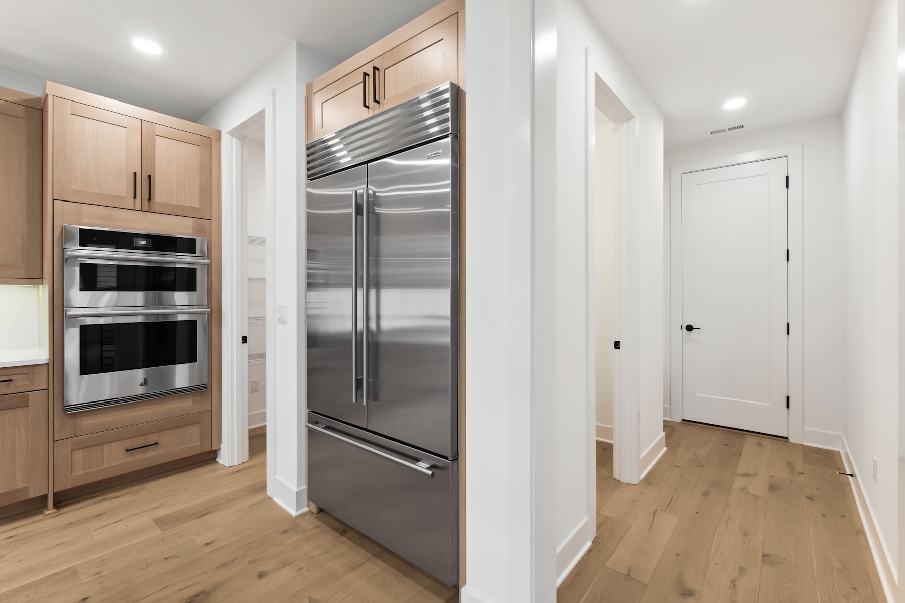 A modern, well-equipped kitchen with stainless steel appliances, light wood cabinetry, and a hardwood floor leading to a hallway.