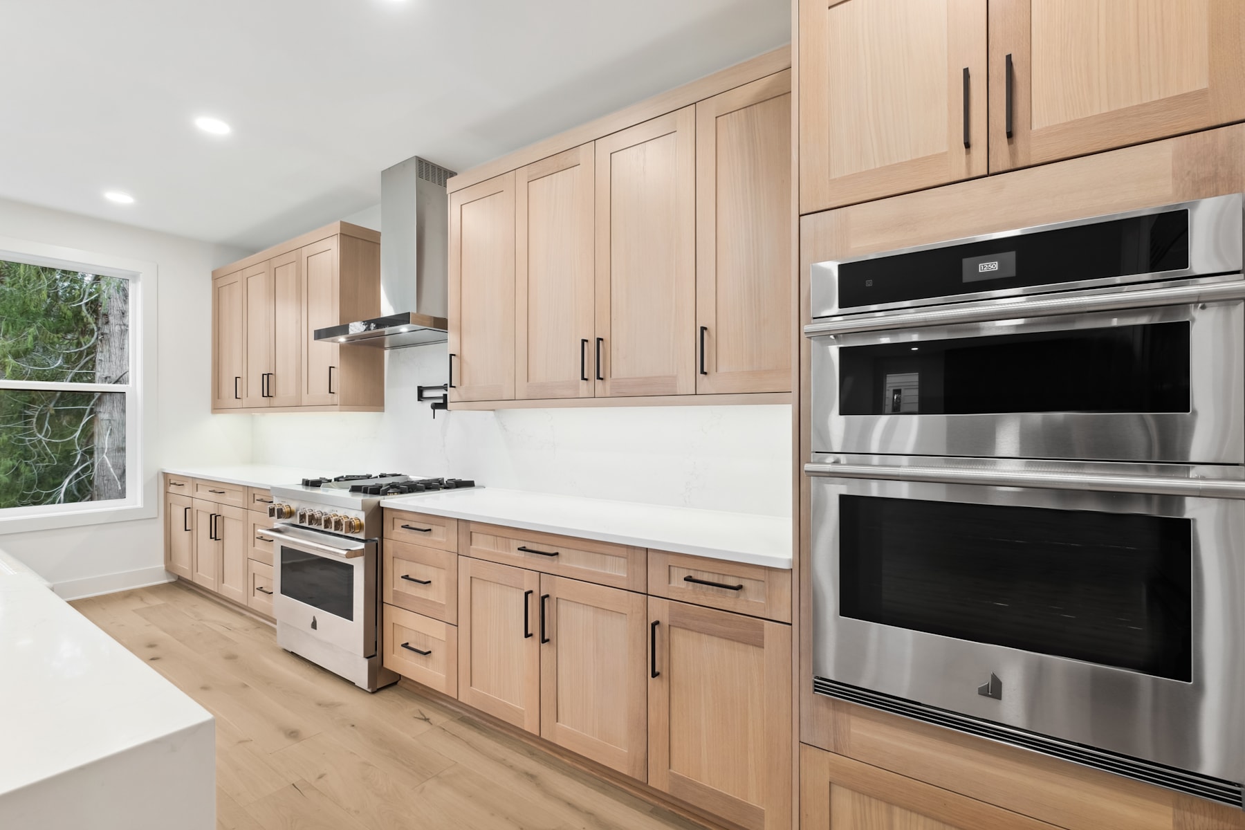A modern, well-equipped kitchen with light-colored wood cabinets, stainless steel appliances, and a large window providing natural light.