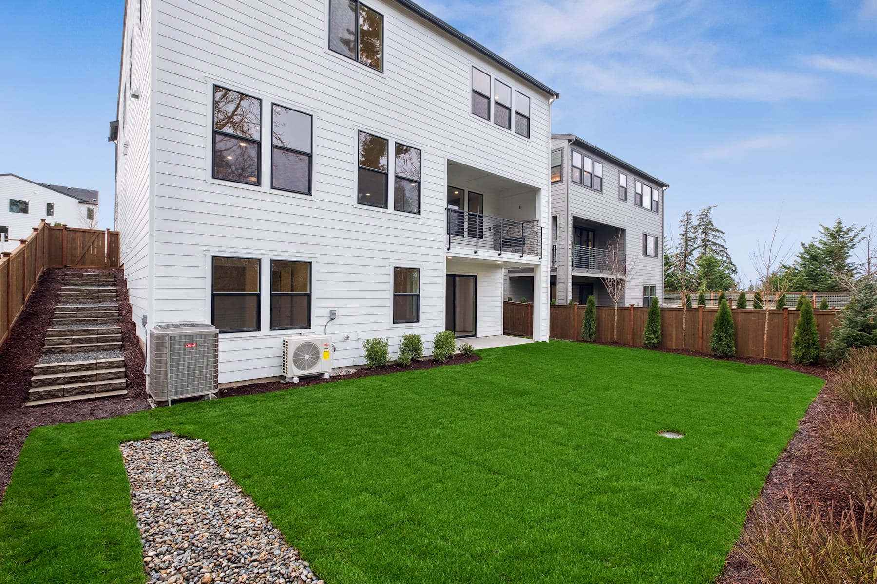A modern, two-story white house with a well-manicured lawn and gravel path in the foreground, surrounded by trees and a clear blue sky in the background.
