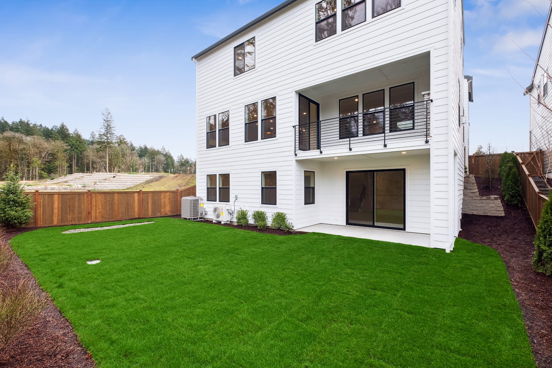 A modern, two-story white house with a well-manicured green lawn in the foreground, surrounded by a wooded area in the background.