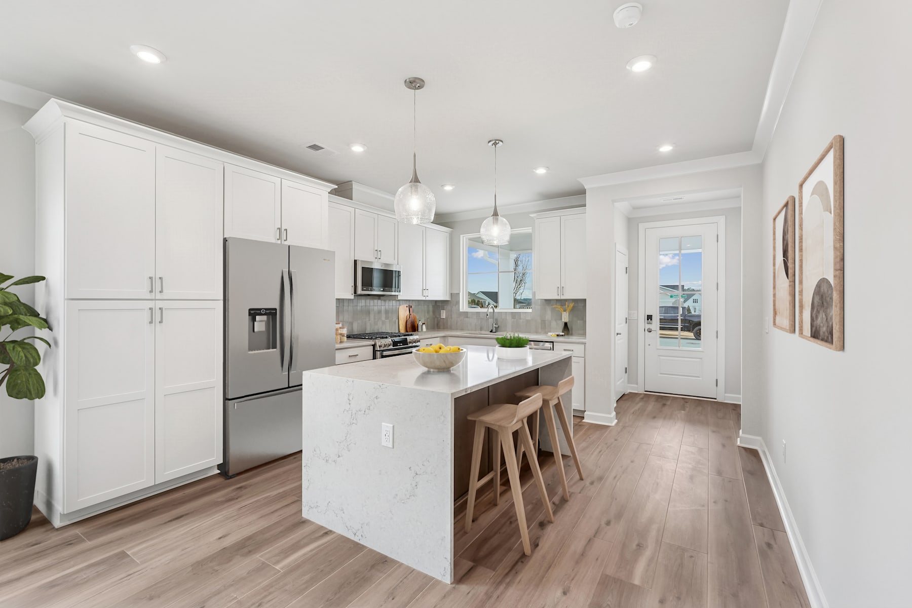 A modern, bright, and spacious kitchen with white cabinets, stainless steel appliances, and a central island with wooden stools, set against a backdrop of hardwood floors and a large window.