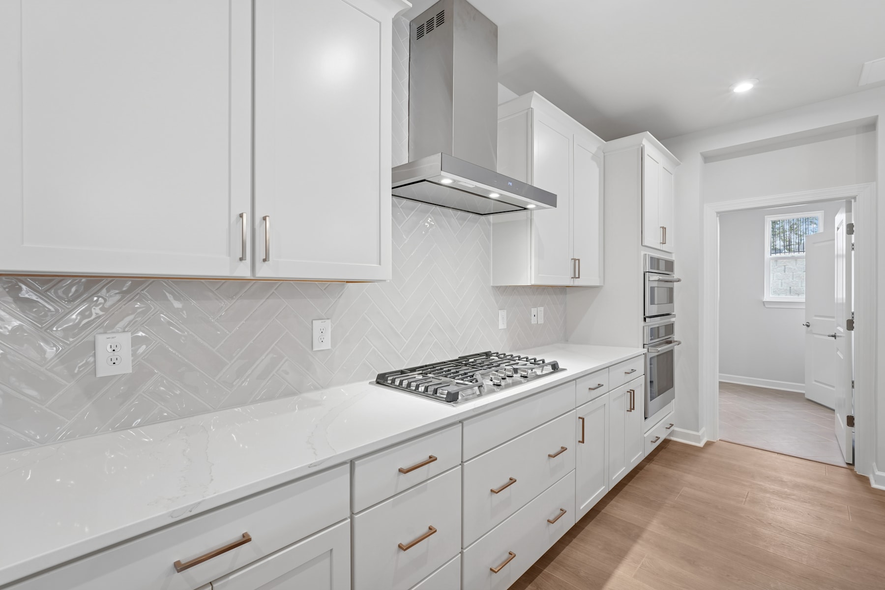 A modern, minimalist kitchen with white cabinets, a stainless steel range hood, and a tiled backsplash, set against a hardwood floor.