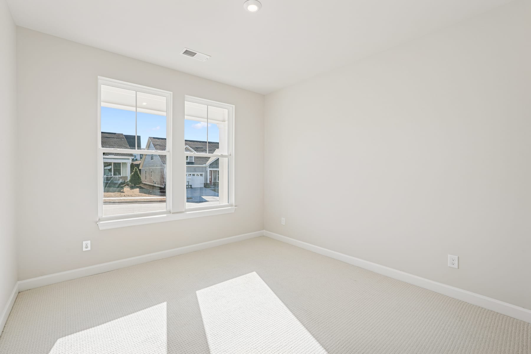 A bright, spacious room with large windows overlooking a residential area, featuring a neutral color scheme and minimal furnishings.