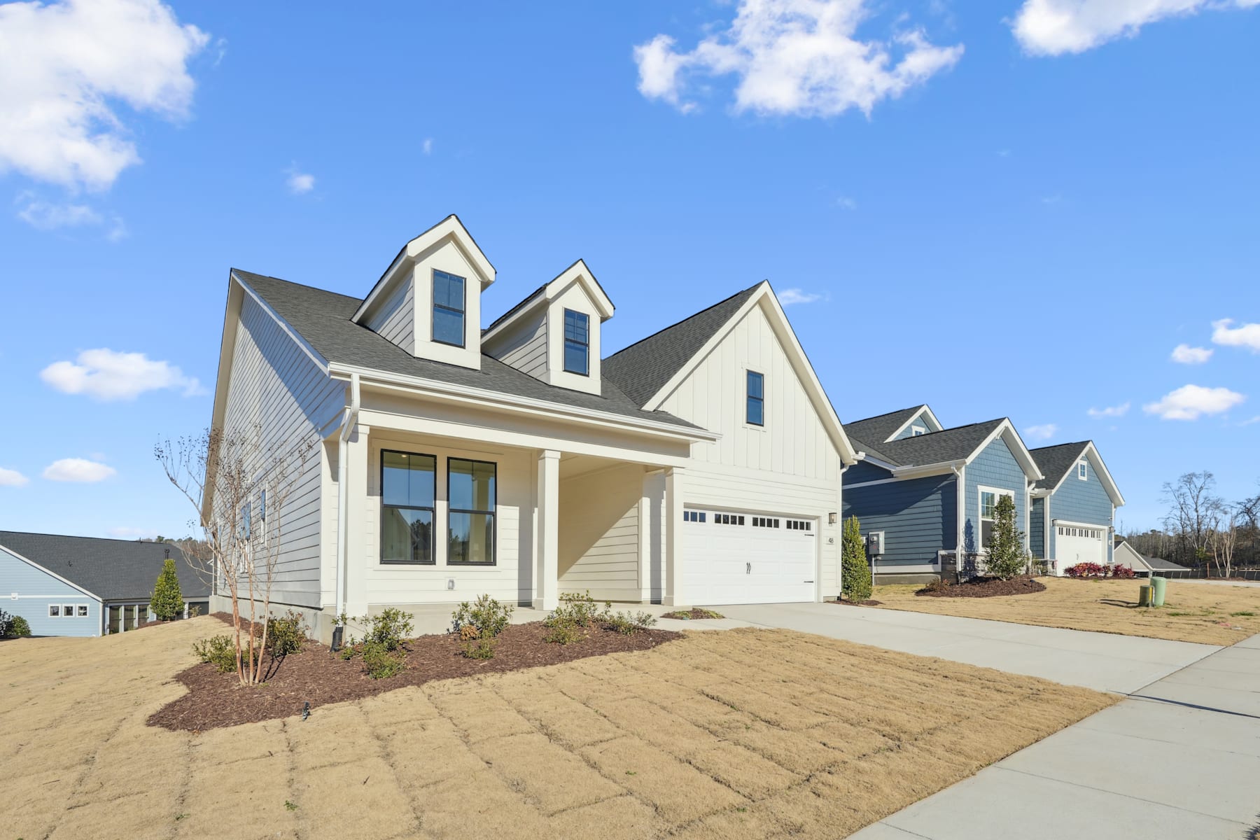 A newly constructed residential neighborhood with a row of beige-colored houses featuring gabled roofs, dormers, and well-manicured landscaping against a backdrop of a blue sky with scattered clouds.