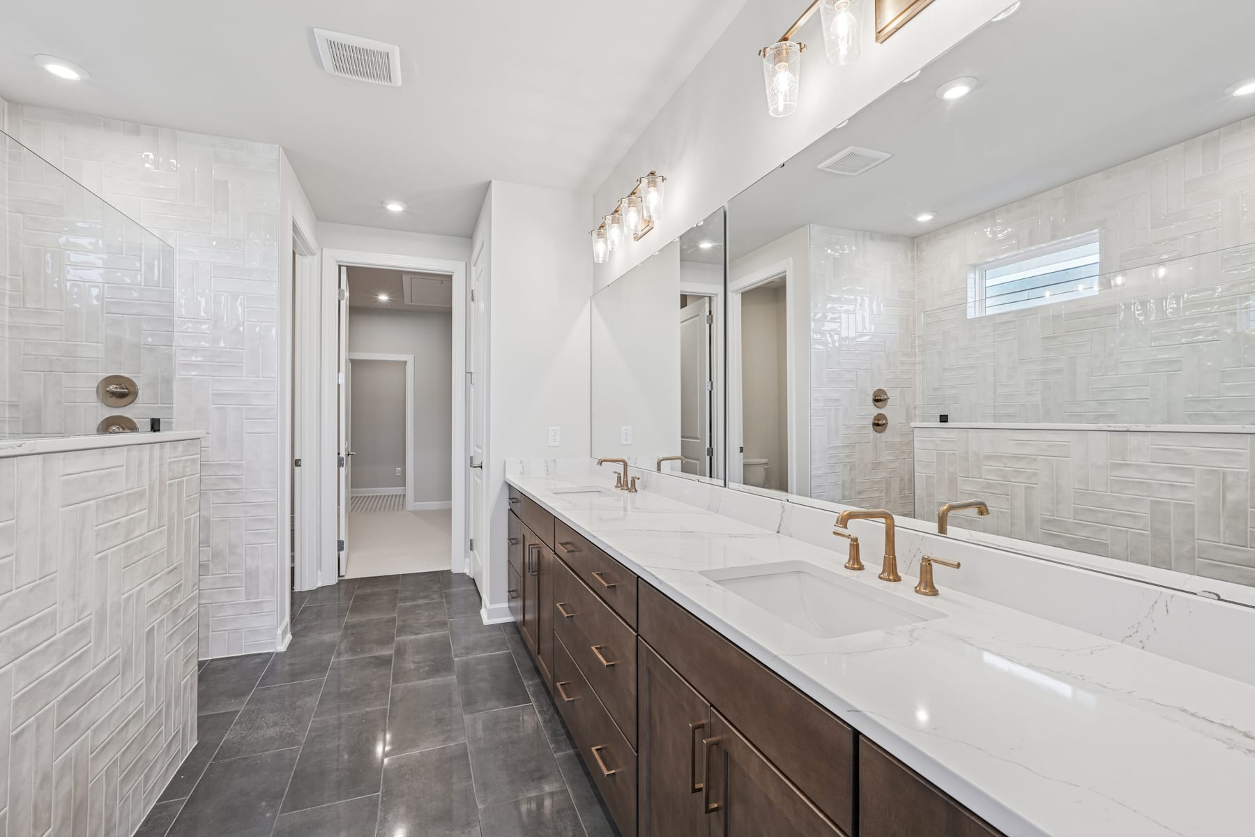 A modern, well-lit bathroom with white tile walls, dark wood vanity, and a double sink with gold fixtures.