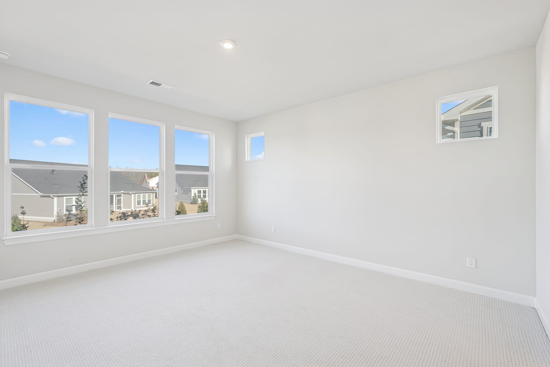 A bright, spacious room with large windows overlooking a residential neighborhood, featuring a clean, minimalist design with white walls and floors.
