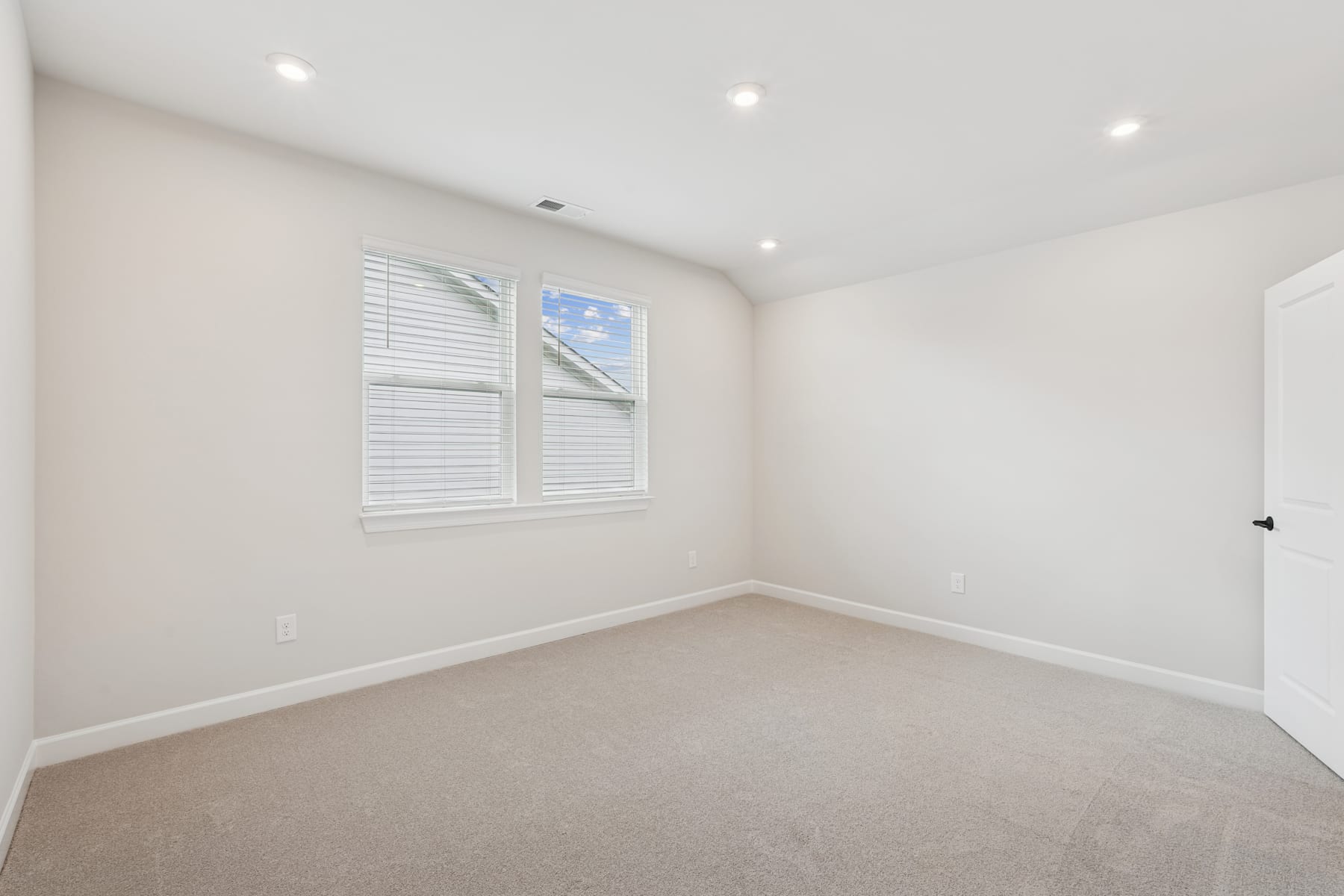 A bright, spacious room with white walls, recessed lighting, and a large window providing natural light. The room appears to be a bedroom or office space with a neutral-colored carpet covering the floor.