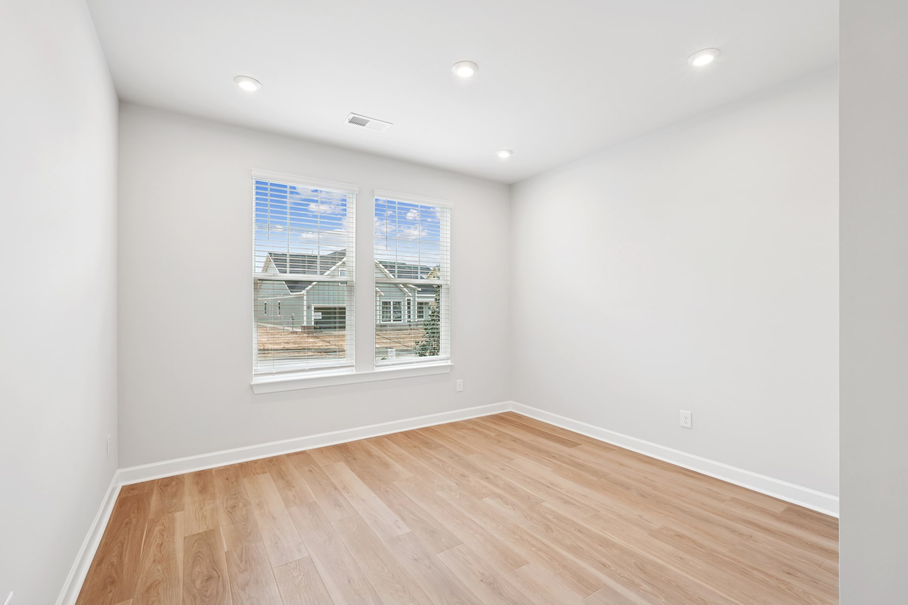 A bright, empty room with white walls, a hardwood floor, and a large window overlooking a residential neighborhood.