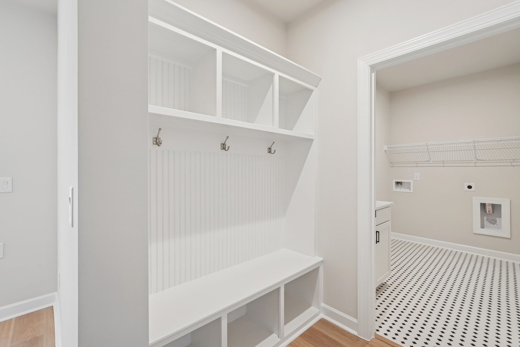 A bright, minimalist mudroom with built-in storage benches and hooks, leading into a tiled bathroom with a checkered floor.