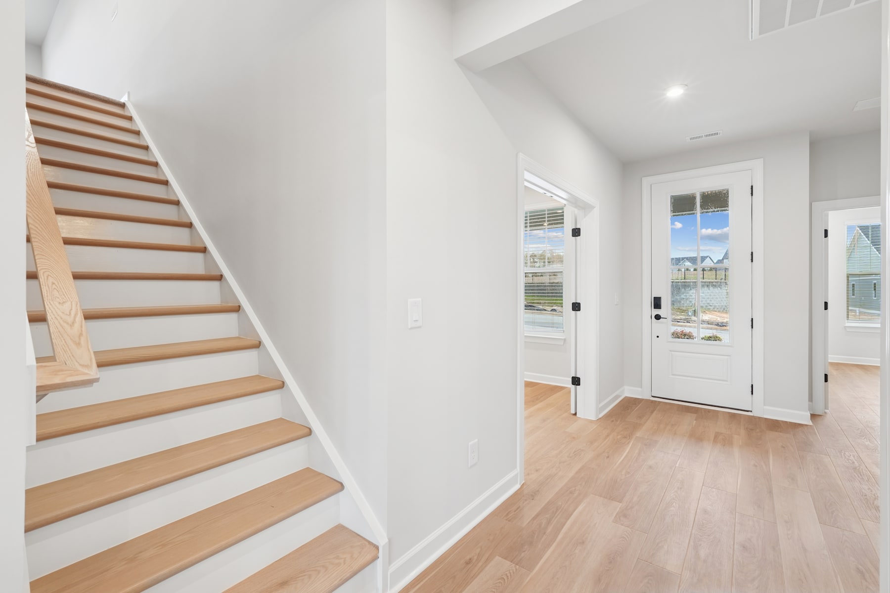 A bright and spacious entryway with a wooden staircase leading upstairs, and a white front door visible in the background, set against a light-colored hardwood floor.