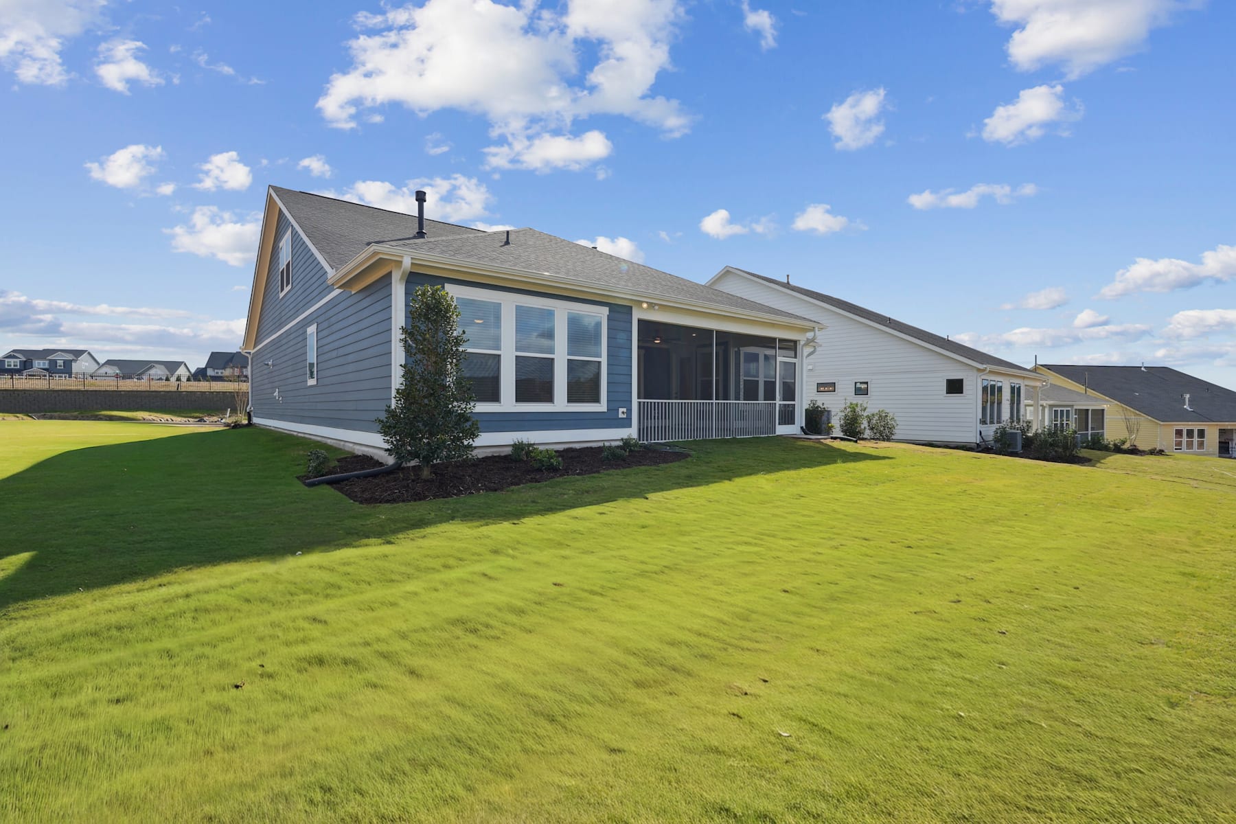 A well-manicured lawn surrounds a modern, two-story house with a gray exterior and large windows, set against a blue sky with fluffy white clouds.