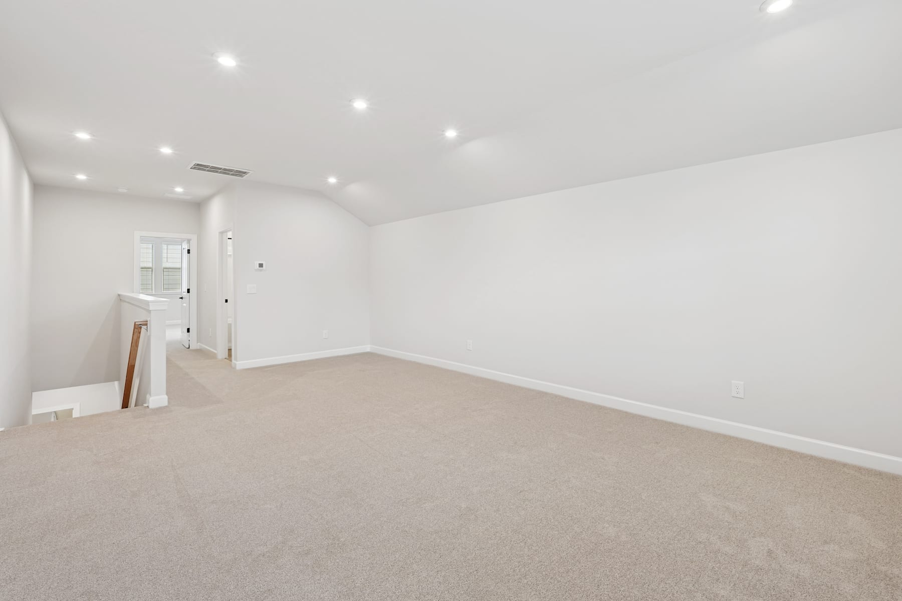 A spacious, well-lit room with white walls and a beige carpeted floor, featuring a doorway leading to another area.