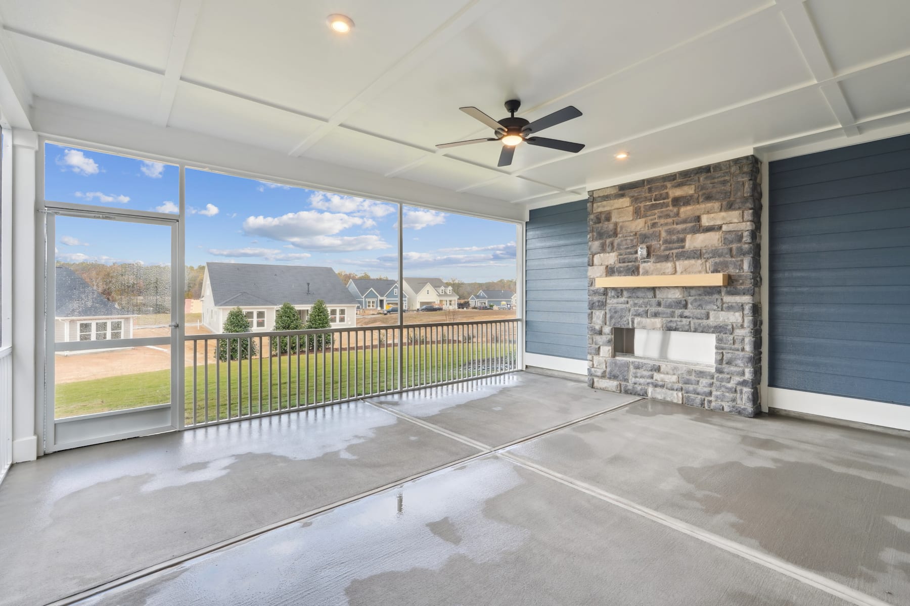 A spacious and bright patio or sunroom with large windows overlooking a residential neighborhood, featuring a stone fireplace, a ceiling fan, and a concrete floor.