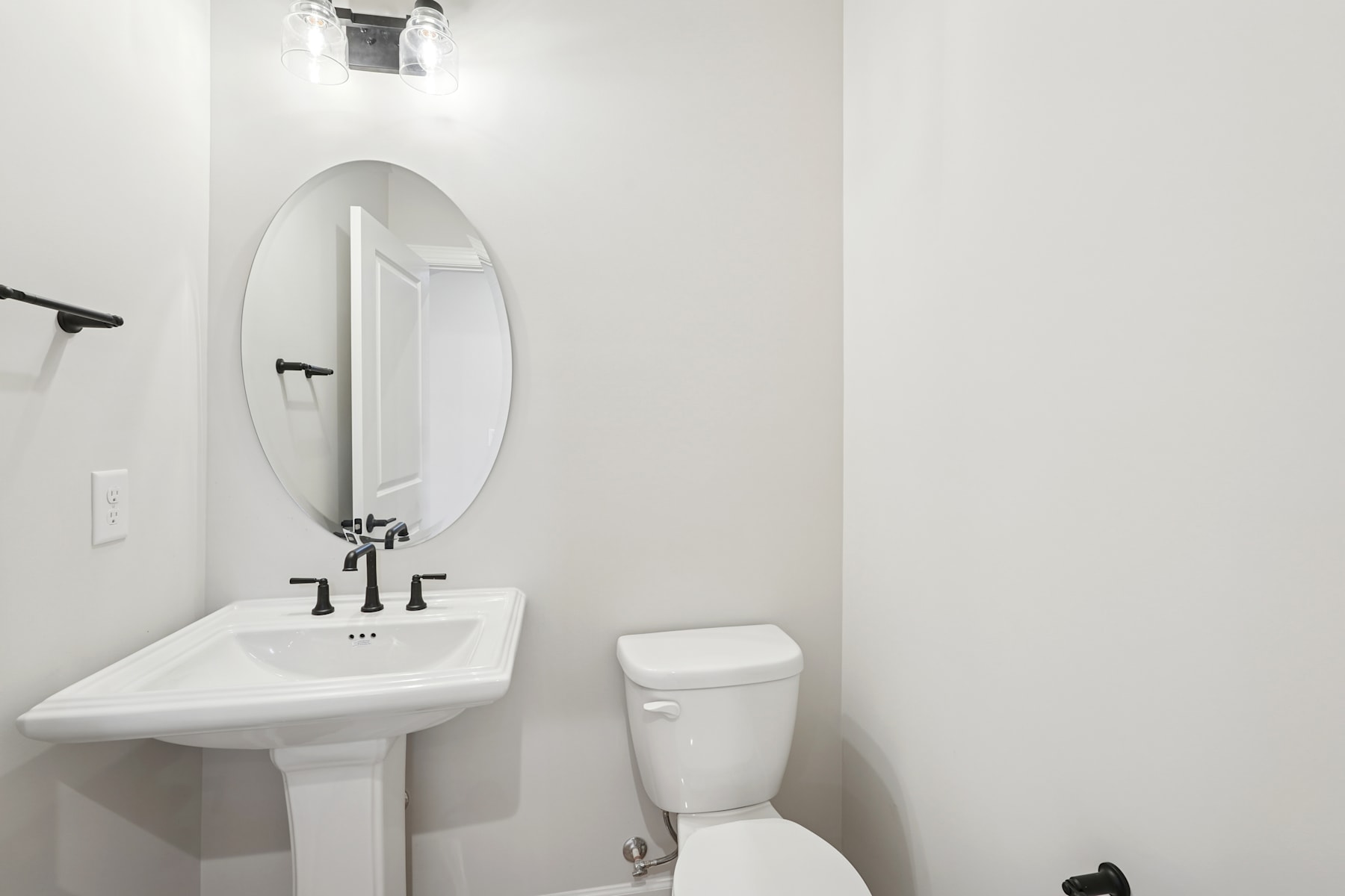 A minimalist bathroom with a white pedestal sink, oval mirror, and a toilet against a plain white wall.