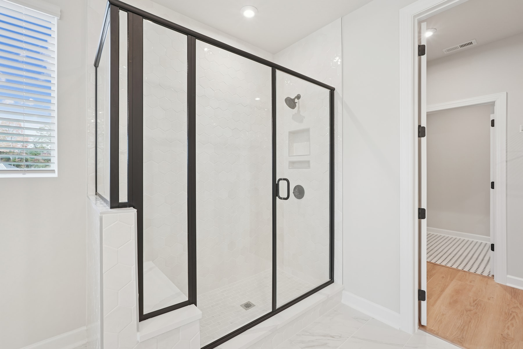 A modern, minimalist bathroom with a large, black-framed glass shower enclosure, a white tiled floor, and a window providing natural light.