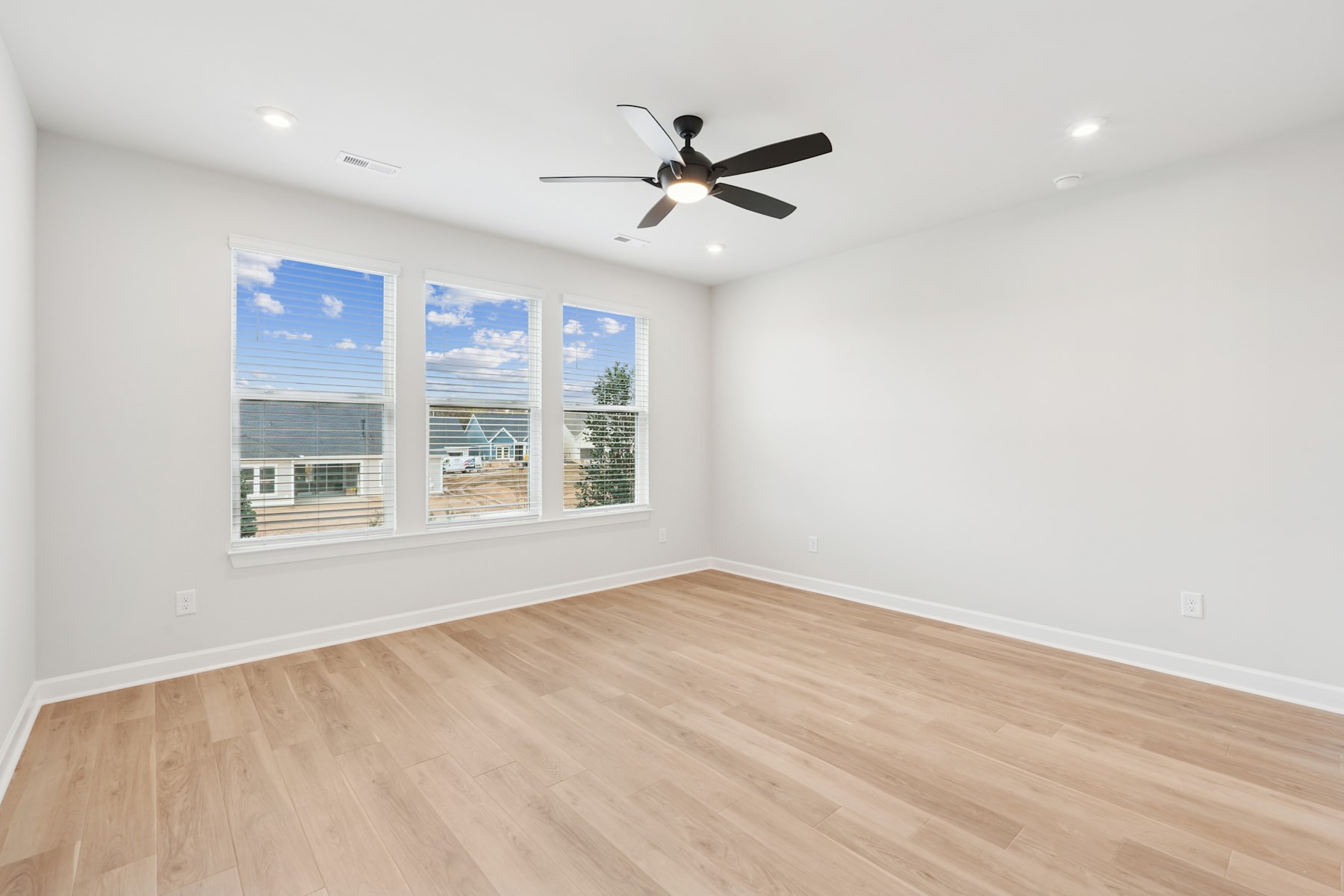 A spacious, well-lit room with a ceiling fan, hardwood floors, and large windows offering a view of the outdoors.