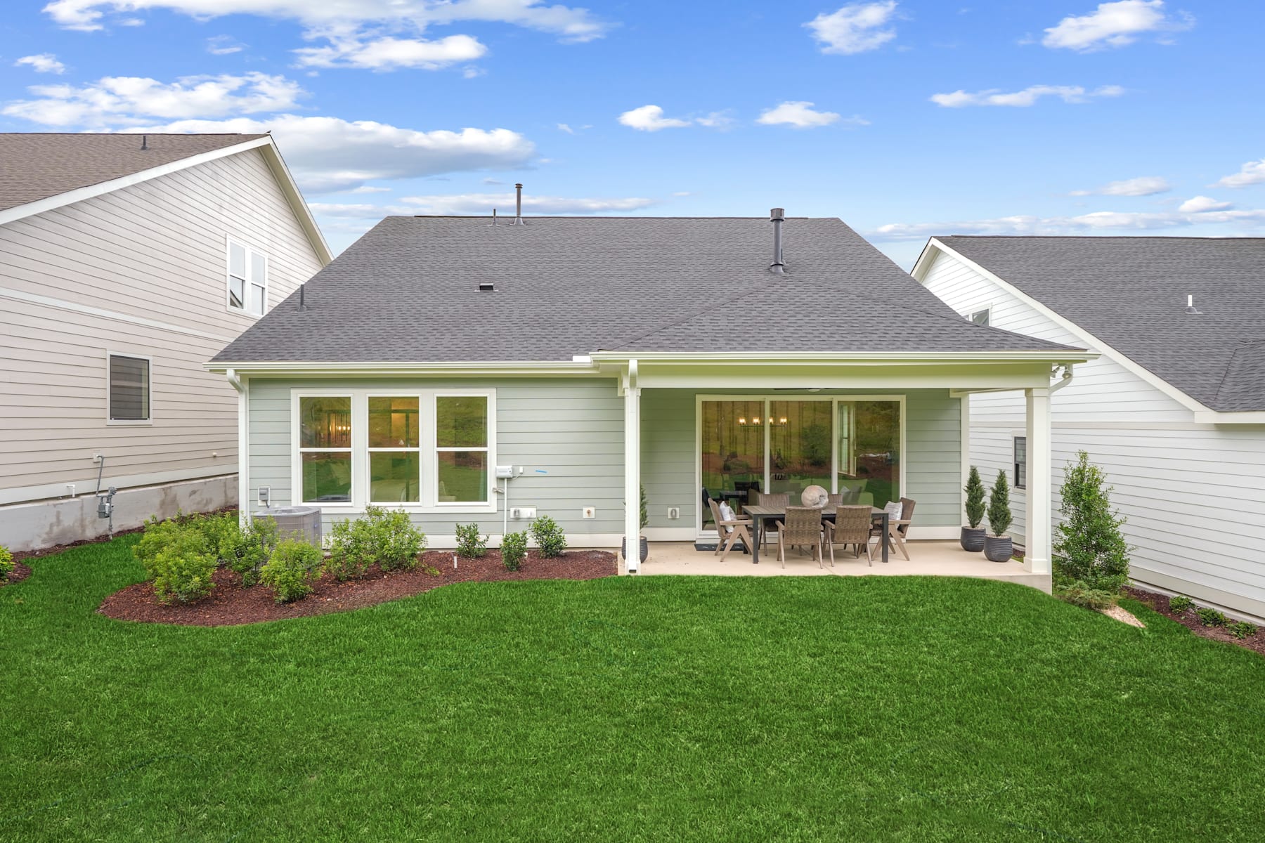 A well-maintained suburban home with a lush green lawn, a covered patio, and a clear blue sky with fluffy white clouds in the background.