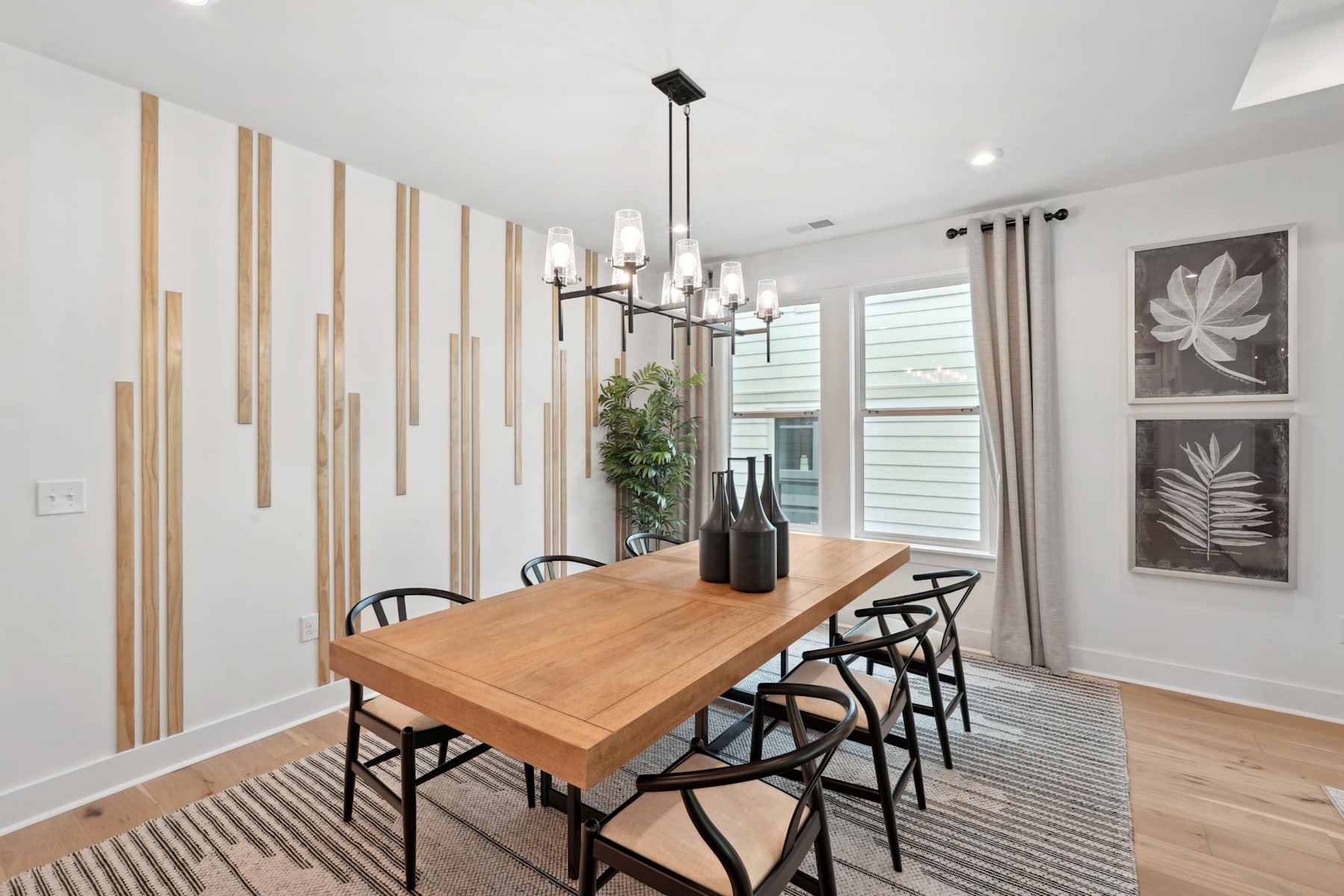 A modern and minimalist dining room with a wooden table, black chairs, and decorative wall panels, complemented by artwork and a chandelier overhead.