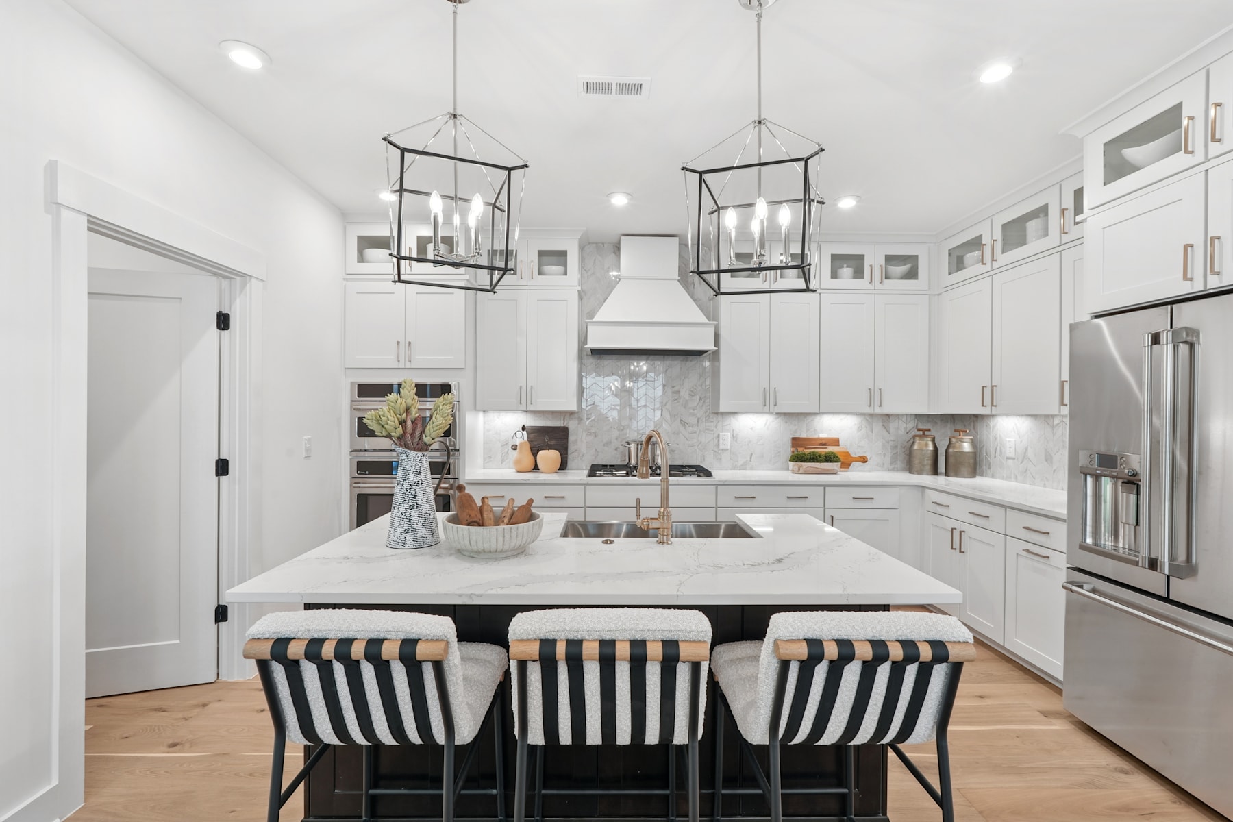 A modern, bright, and spacious kitchen with white cabinets, a large island with striped stools, and pendant lighting fixtures.