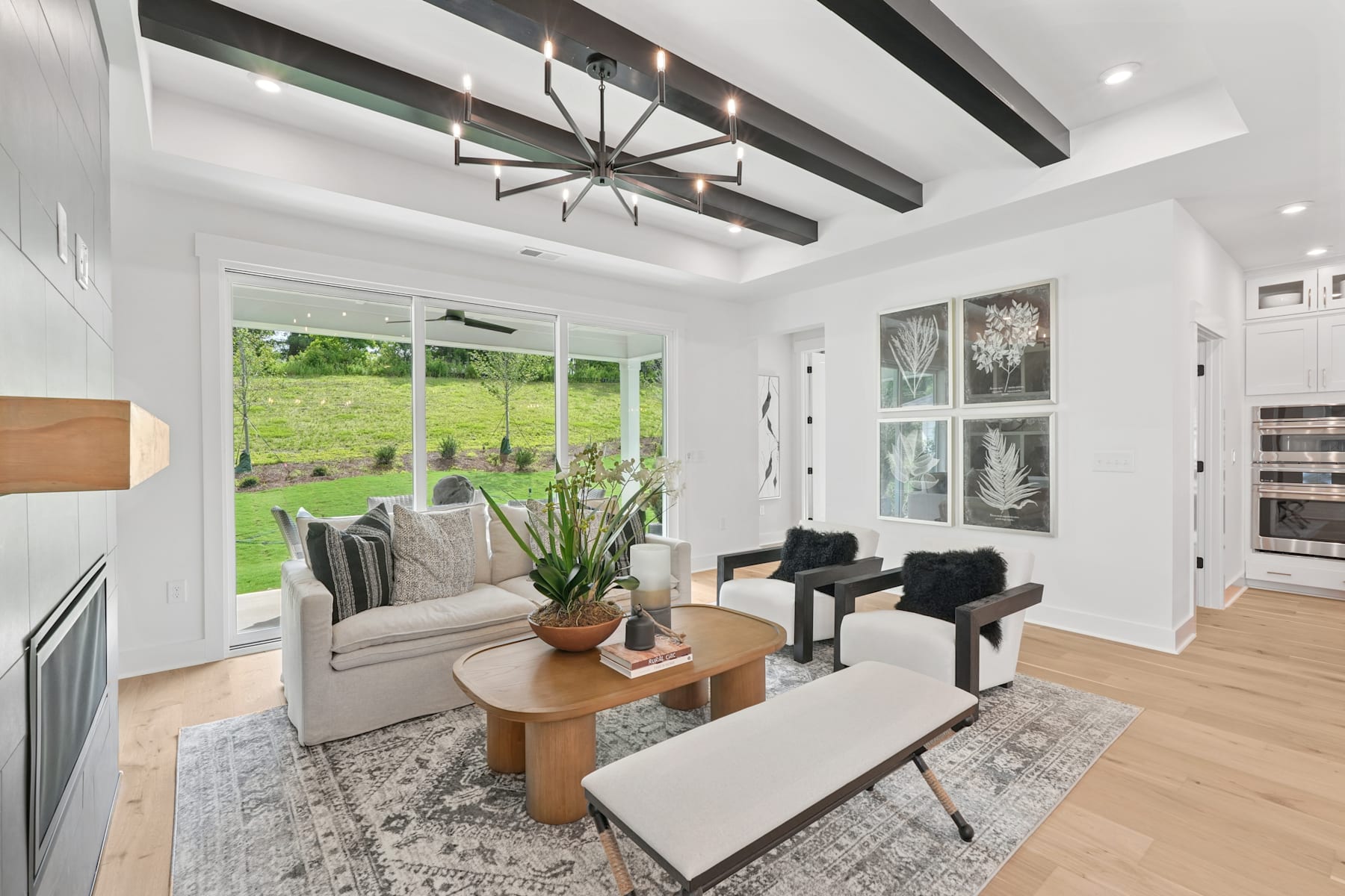 A spacious and bright living room with large windows overlooking a lush green outdoor area, featuring a cozy seating arrangement, a modern chandelier, and a wooden coffee table on a patterned rug.