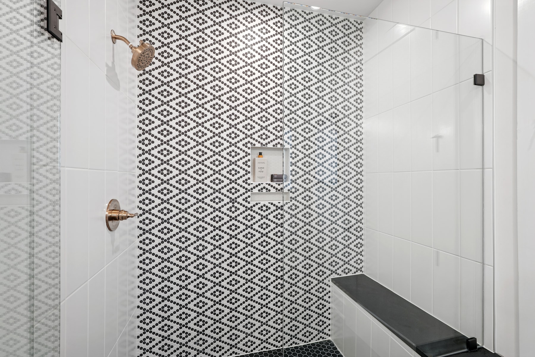 The image shows a modern bathroom with a patterned tile wall in a black and white diamond design, a glass shower enclosure, and a black bench or shelf in the foreground.