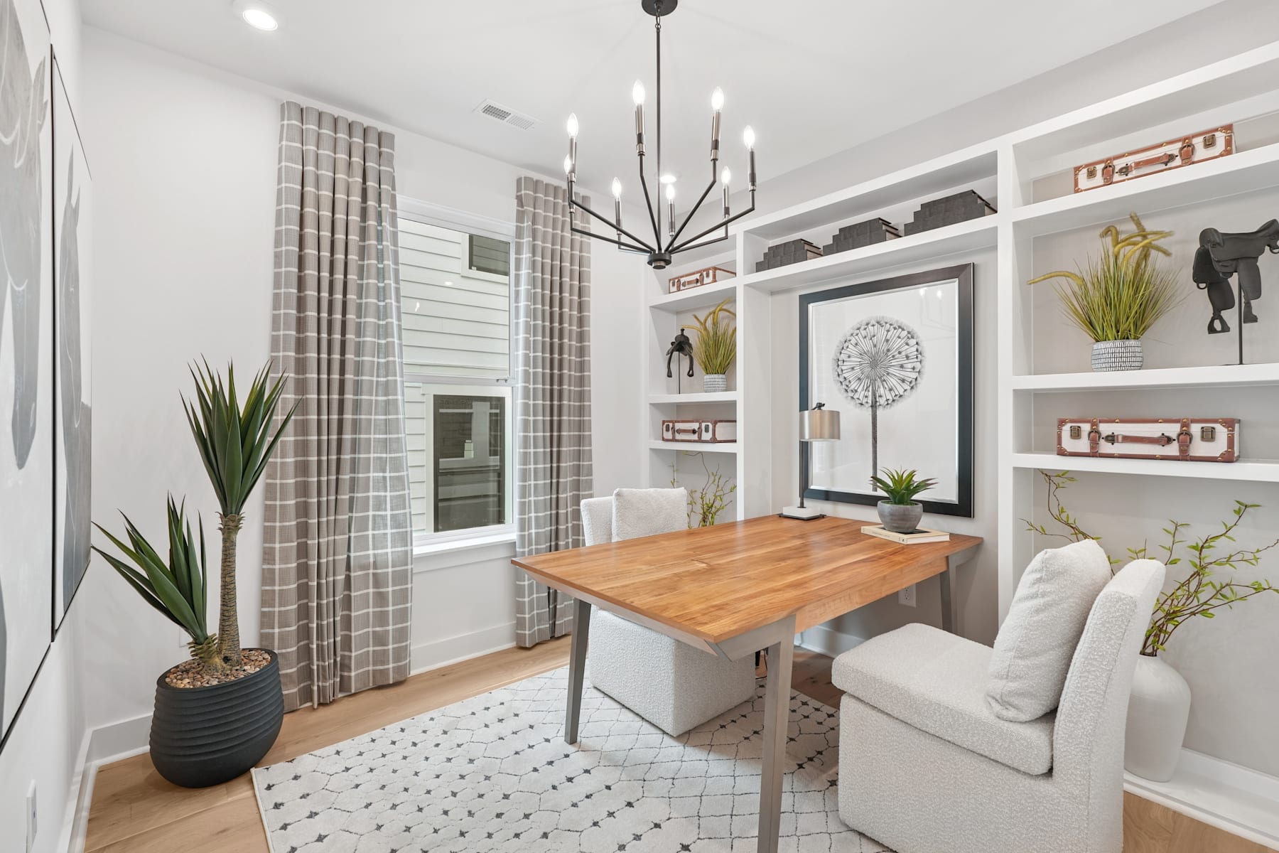 A cozy and well-designed living room with a wooden dining table, a comfortable sofa, and various decorative elements such as plants, artwork, and a modern chandelier.