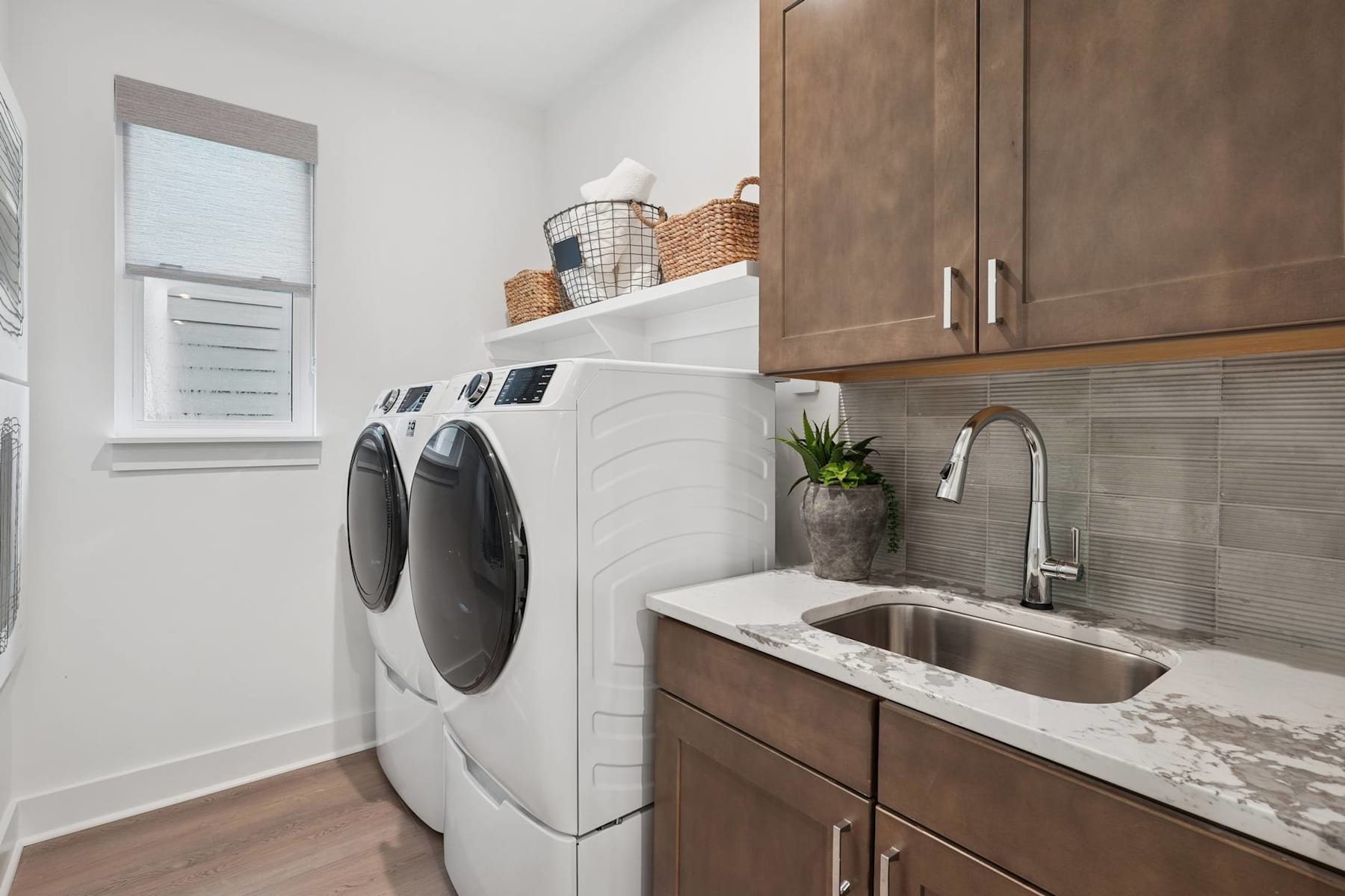 A modern and well-organized laundry room with a washer and dryer, wooden cabinets, and a sink with a marble countertop, along with various storage baskets and a potted plant.