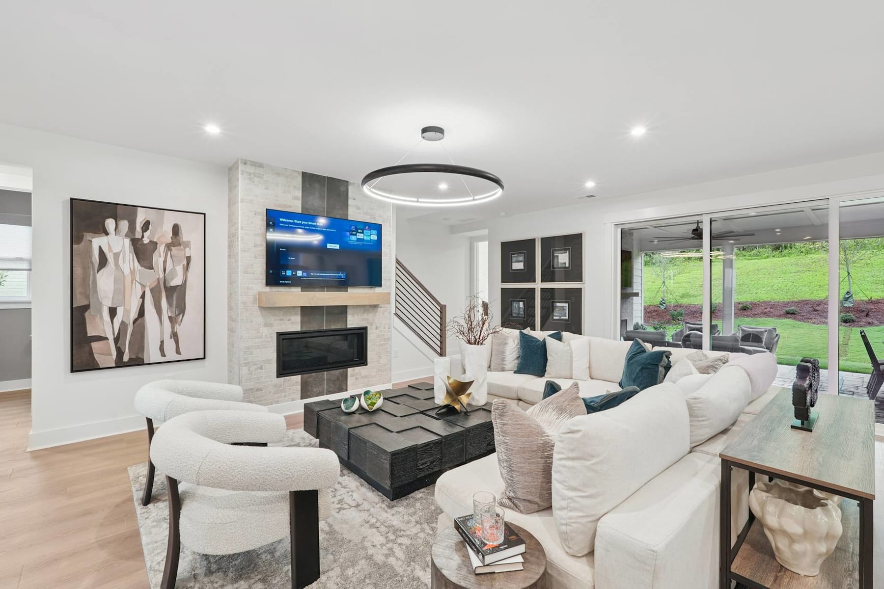 A modern and spacious living room with a large sectional sofa, a fireplace, and a flat-screen TV mounted on the wall. The room features hardwood floors, large windows, and various decorative elements.