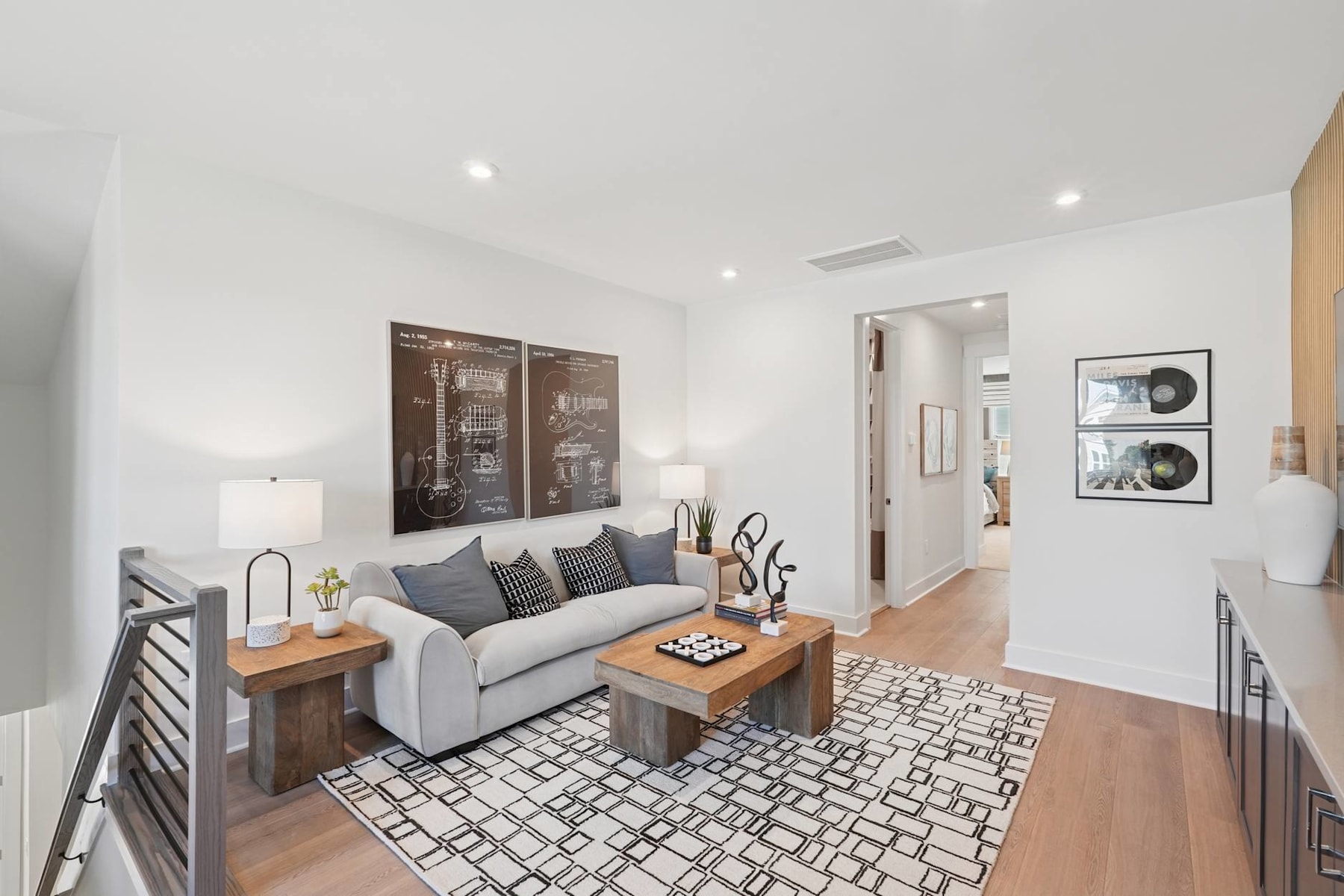 A modern and minimalist living room with a gray sofa, wooden coffee table, and a patterned area rug on the hardwood floor. The room features white walls, recessed lighting, and framed artwork on the walls.