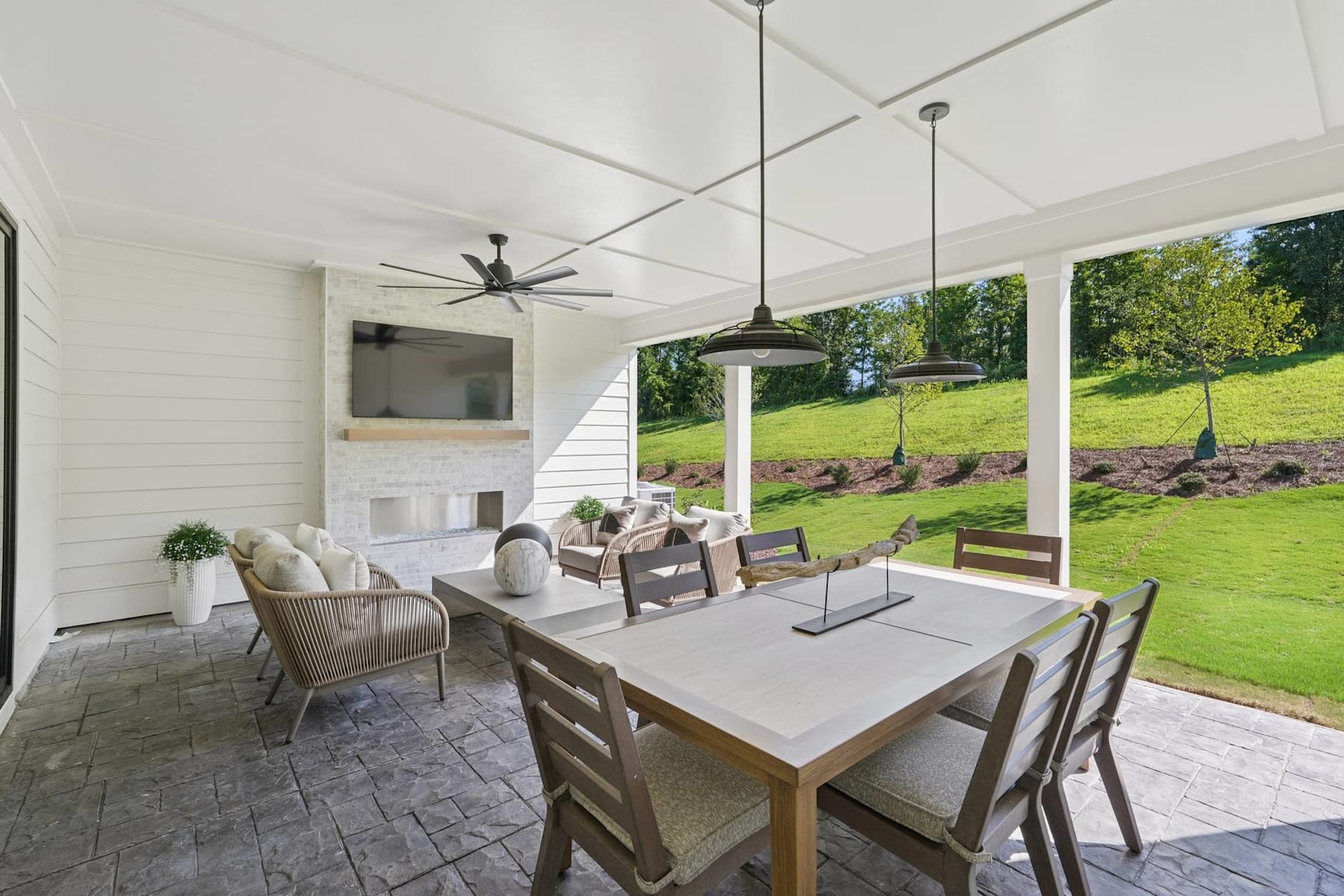 A cozy and inviting indoor-outdoor dining area with a wooden table, comfortable chairs, and a scenic view of a lush green lawn and trees through the large windows.