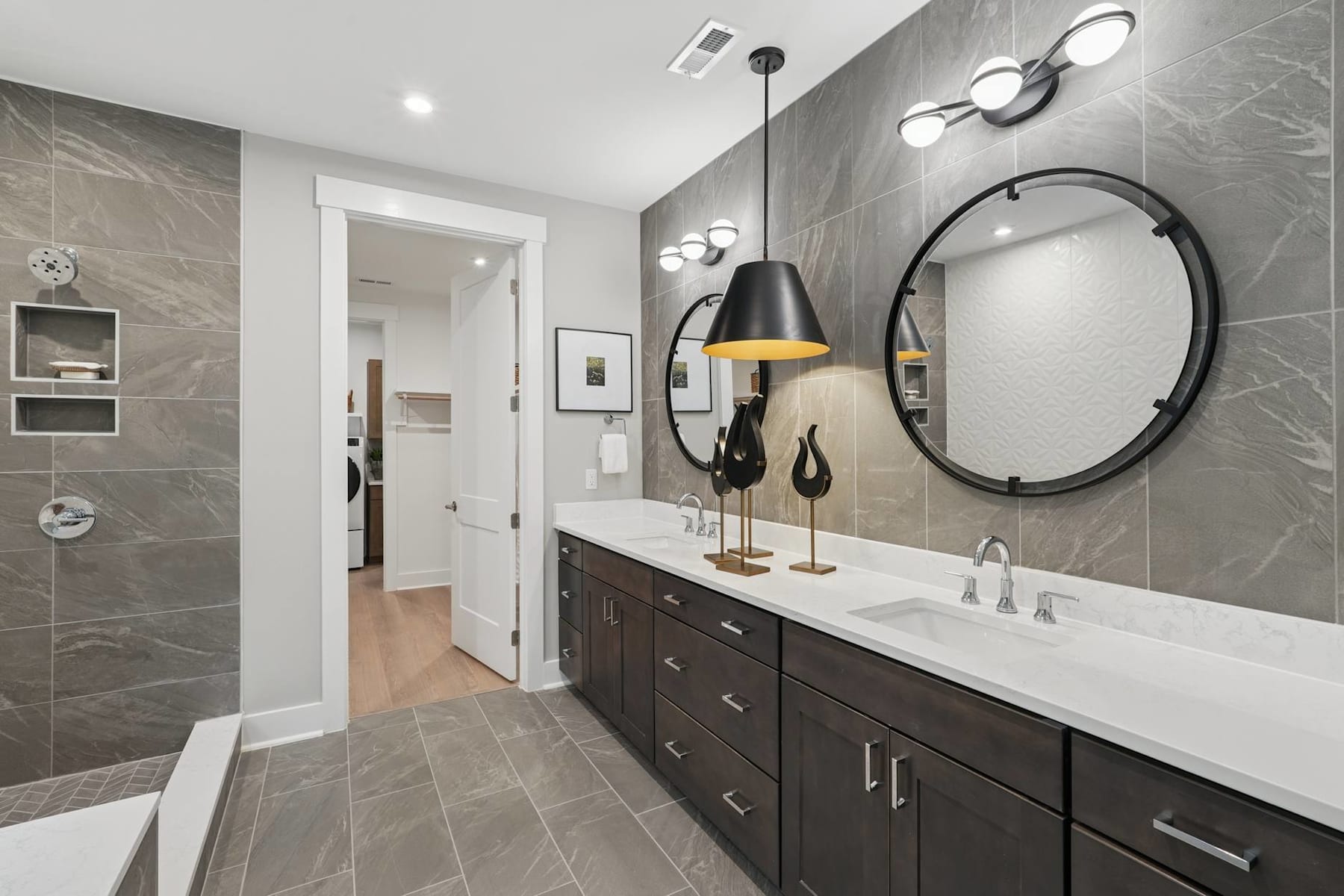 A modern and stylish bathroom with a large vanity, a round mirror, and various lighting fixtures, set against a backdrop of gray tiled walls.