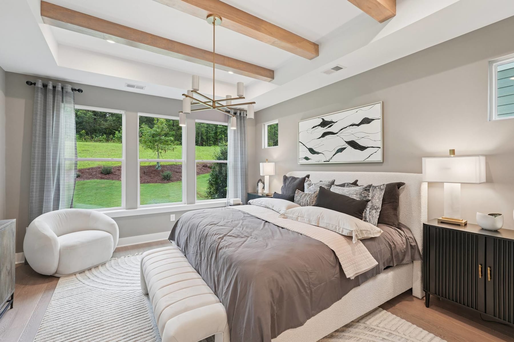 A cozy and inviting bedroom with a large bed, plush bedding, and a modern chandelier overhead. The room features wooden beams on the ceiling, large windows overlooking a lush outdoor landscape, and a stylish artwork on the wall.