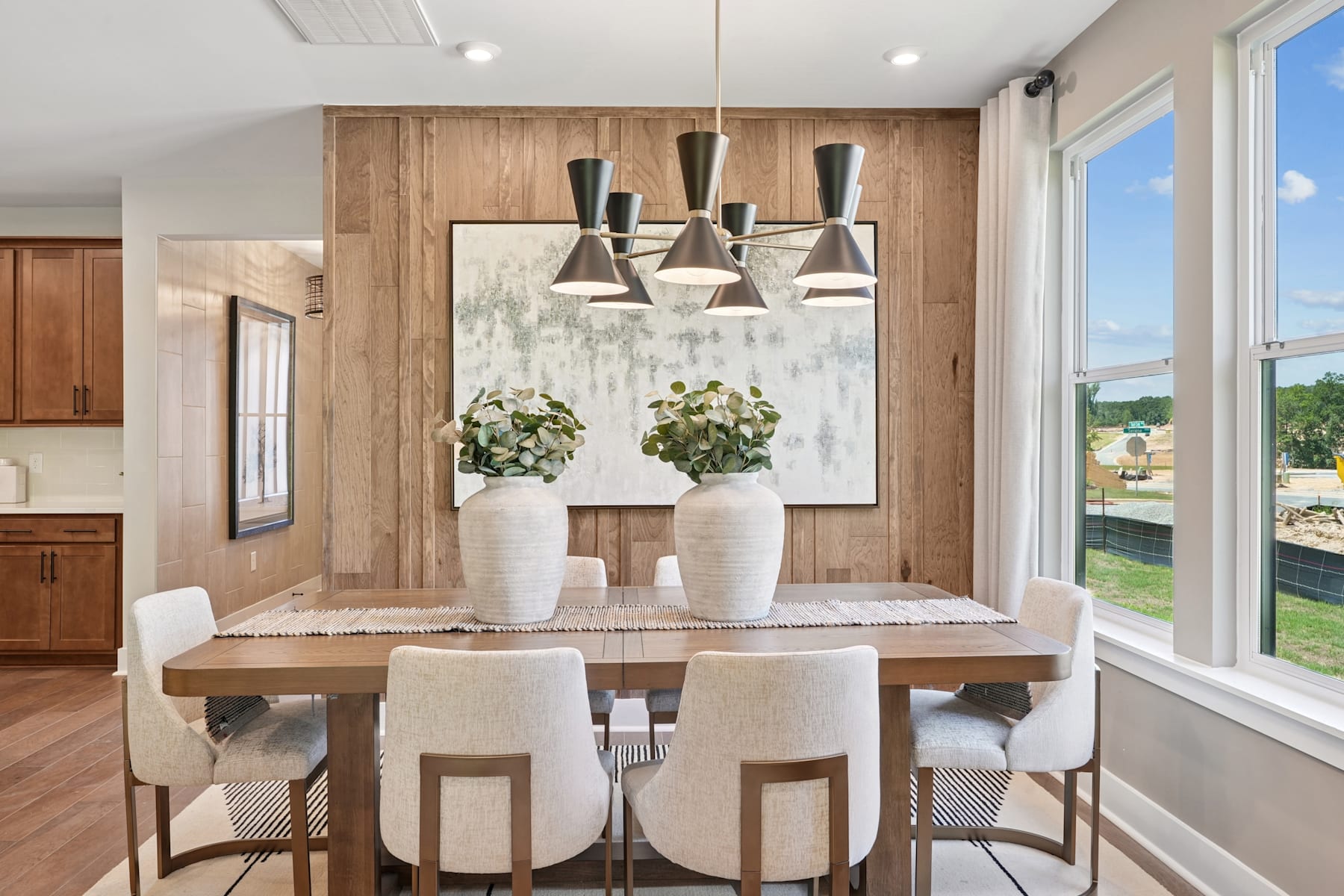 A modern and cozy dining room with a wooden accent wall, a large window overlooking the outdoors, and a stylish pendant lighting fixture above the wooden dining table and chairs.