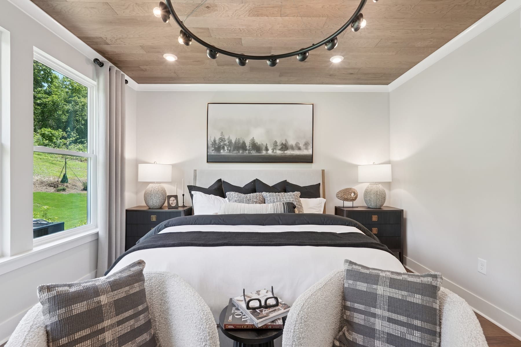 A cozy and inviting bedroom with a large window overlooking a lush, green landscape, featuring a plush bed with patterned bedding and decorative pillows, complemented by a rustic-style lighting fixture overhead.