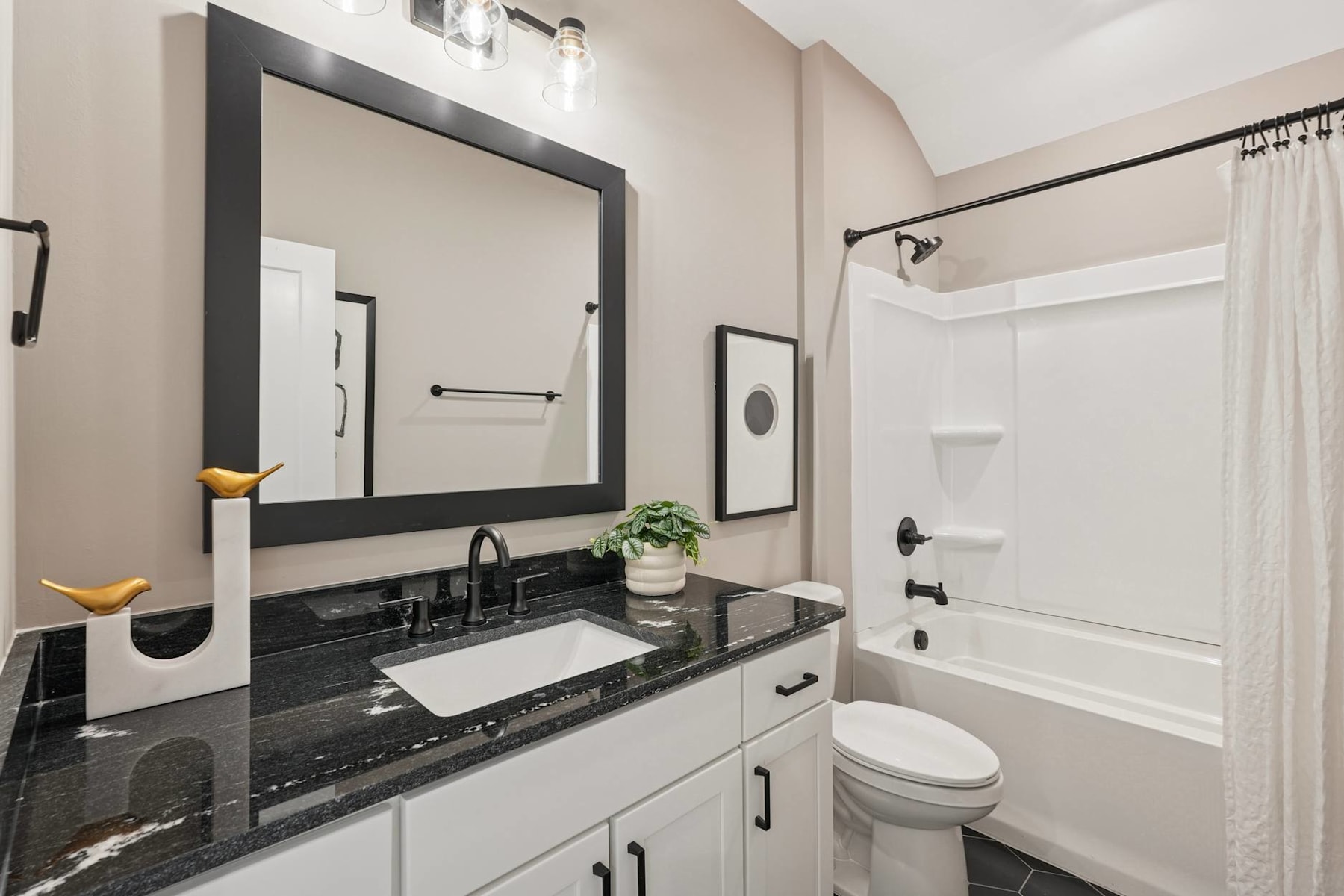 A modern and stylish bathroom with a large mirror, a white vanity with a black granite countertop, and a shower enclosure in the background.