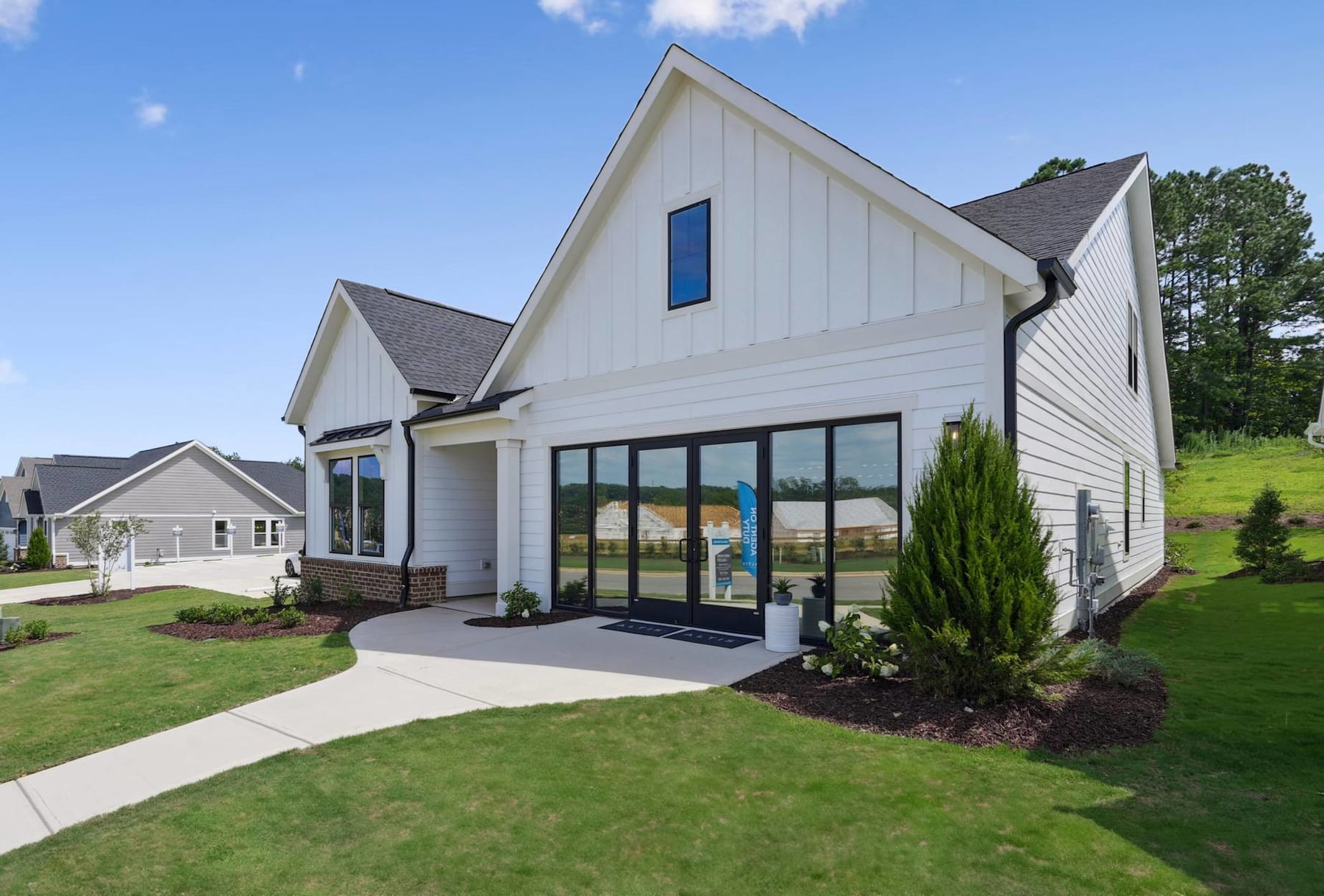 A modern, two-story white house with a slanted roof and large windows stands in a well-manicured yard with lush greenery and a paved walkway leading to the entrance.