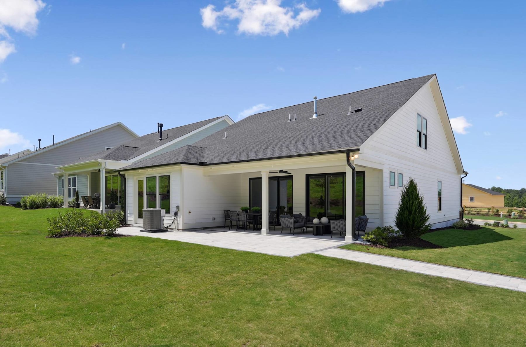 A modern, two-story house with a covered patio and a well-manicured lawn in the foreground, set against a clear blue sky with scattered clouds.