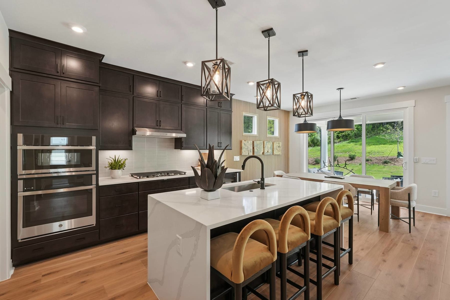 A modern and spacious kitchen with dark wood cabinets, a white island countertop, and pendant lighting fixtures, overlooking a scenic outdoor view through large windows.