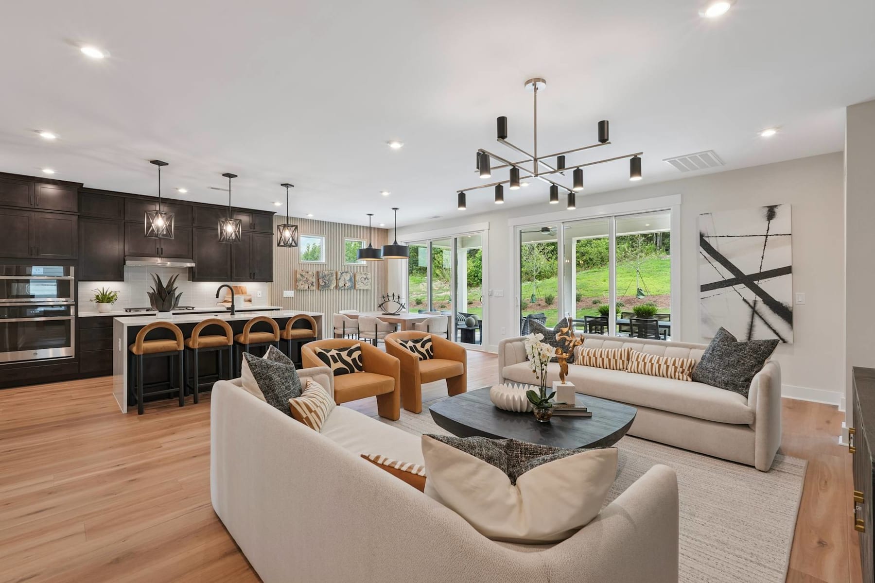 A modern and spacious open-concept living room and kitchen with dark wood cabinetry, a large chandelier, and comfortable seating arrangements, all surrounded by large windows overlooking a lush outdoor landscape.