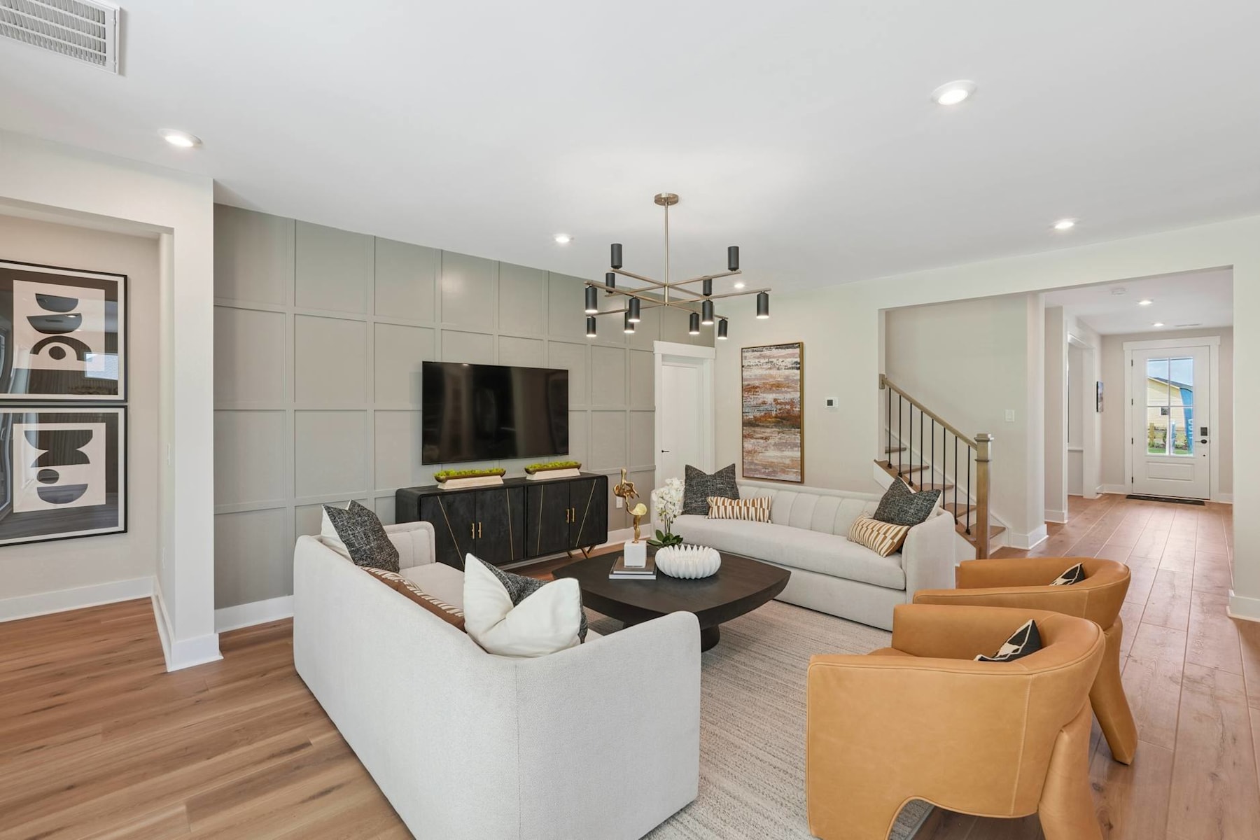 A modern and stylish living room with a comfortable seating arrangement, a large TV, and a chandelier overhead, set against a neutral color palette and hardwood flooring.