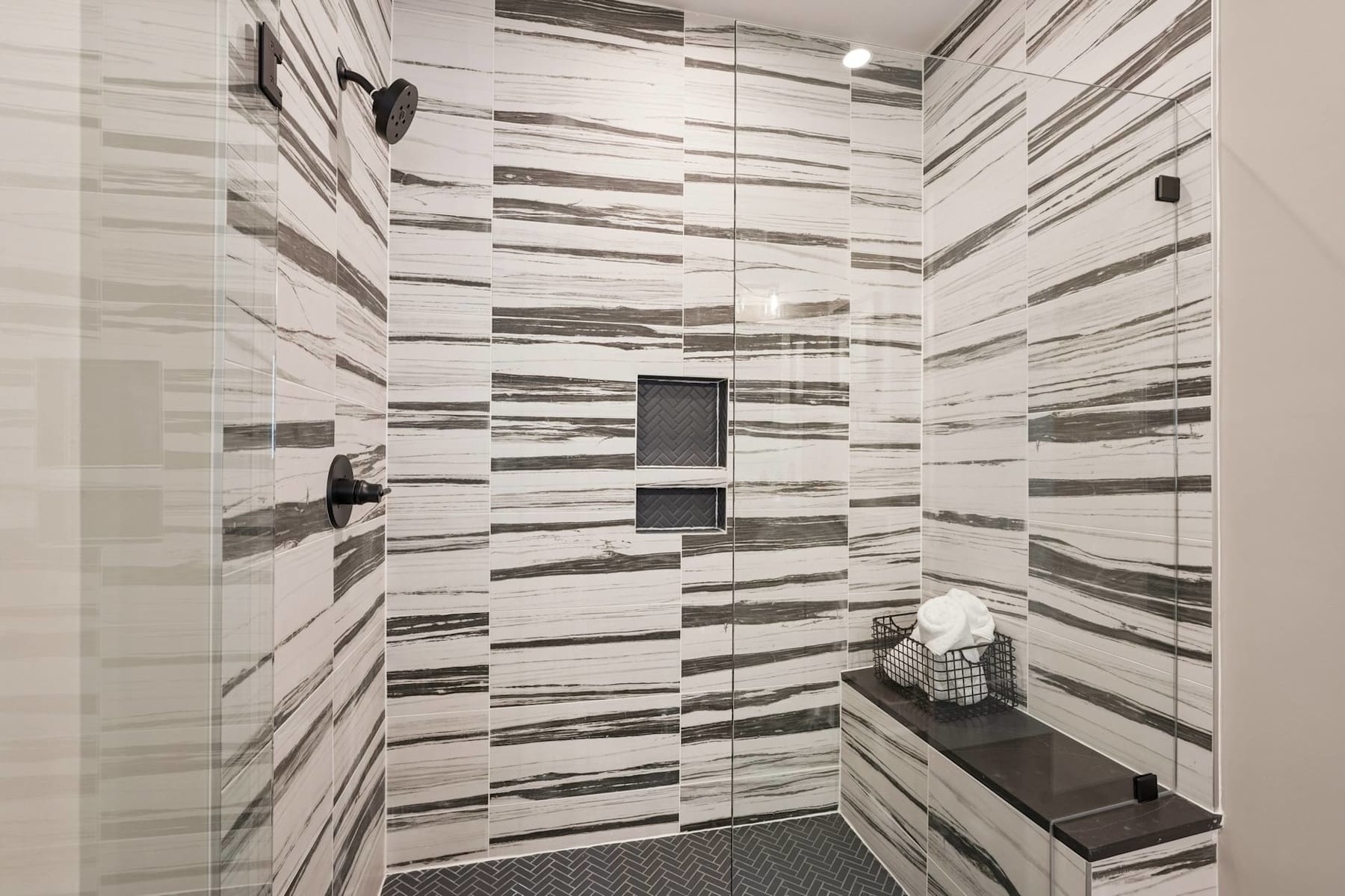 The image depicts a bathroom interior with a tiled wall pattern featuring alternating horizontal stripes of varying shades of gray. The foreground includes a recessed niche or shelf, and a towel or cloth can be seen on the floor.