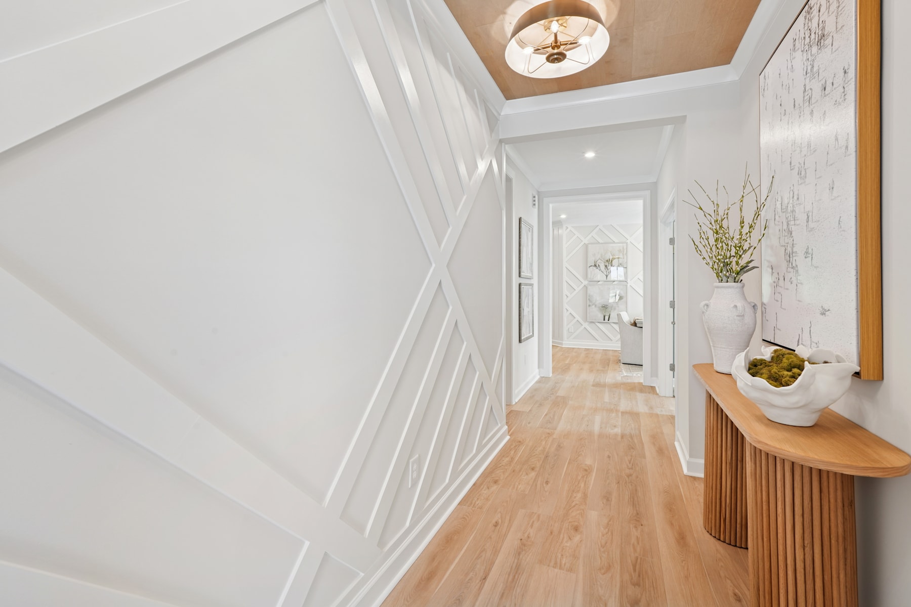 A bright and airy hallway with white walls, wooden floors, and a decorative light fixture overhead, leading to a cozy-looking space with a wooden console table and floral decor.