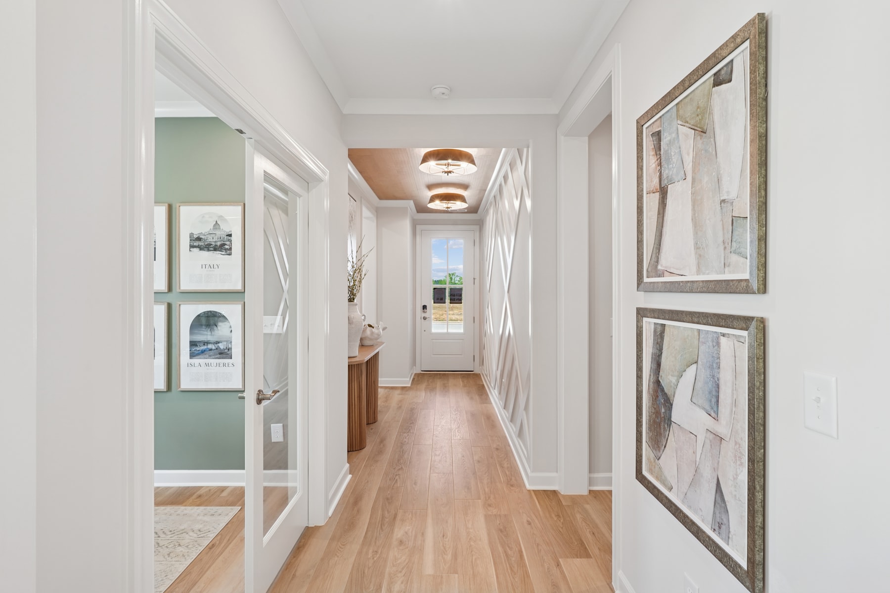 A bright and airy hallway with wooden floors, white walls, and framed artwork hanging on the walls, leading to a doorway at the end.