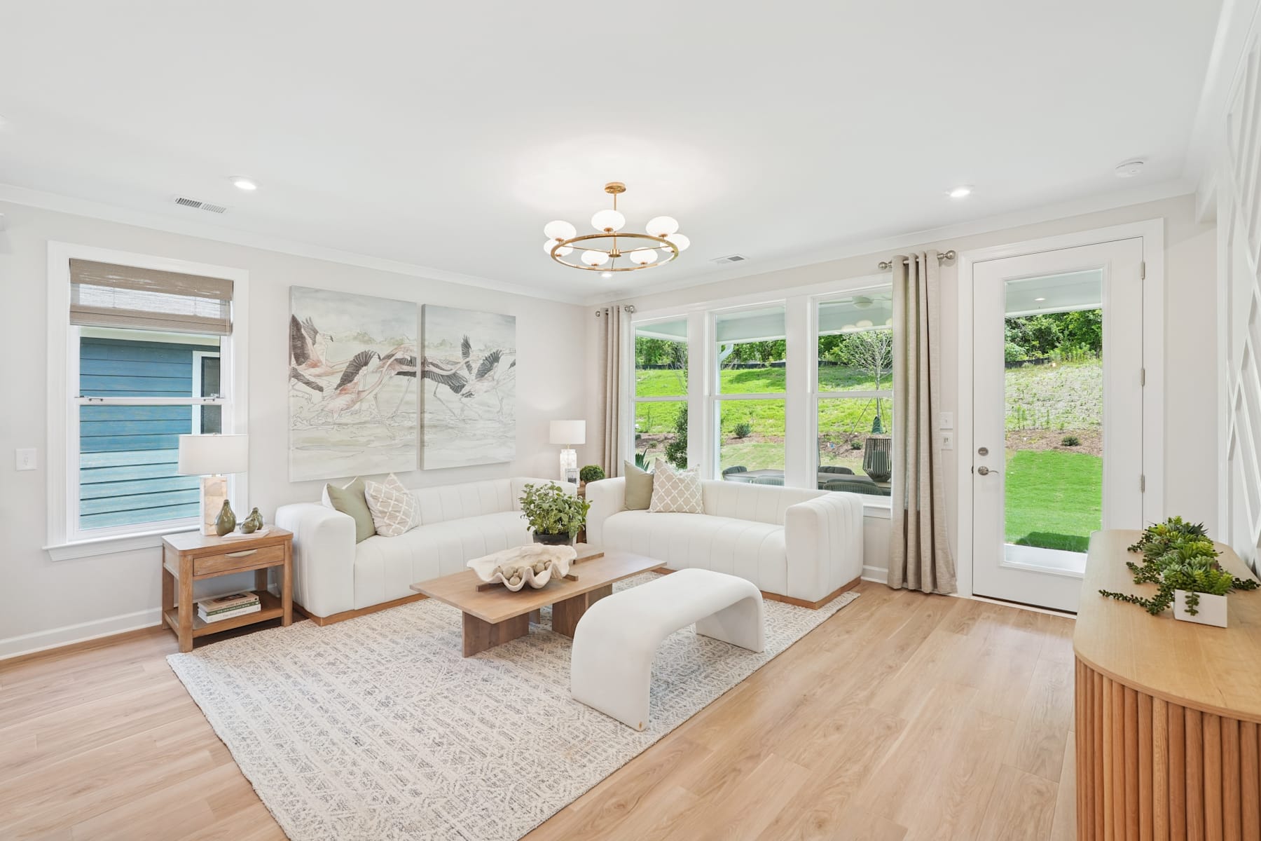 A bright and airy living room with white furniture, a large window overlooking a lush green backyard, and a decorative wall art piece.