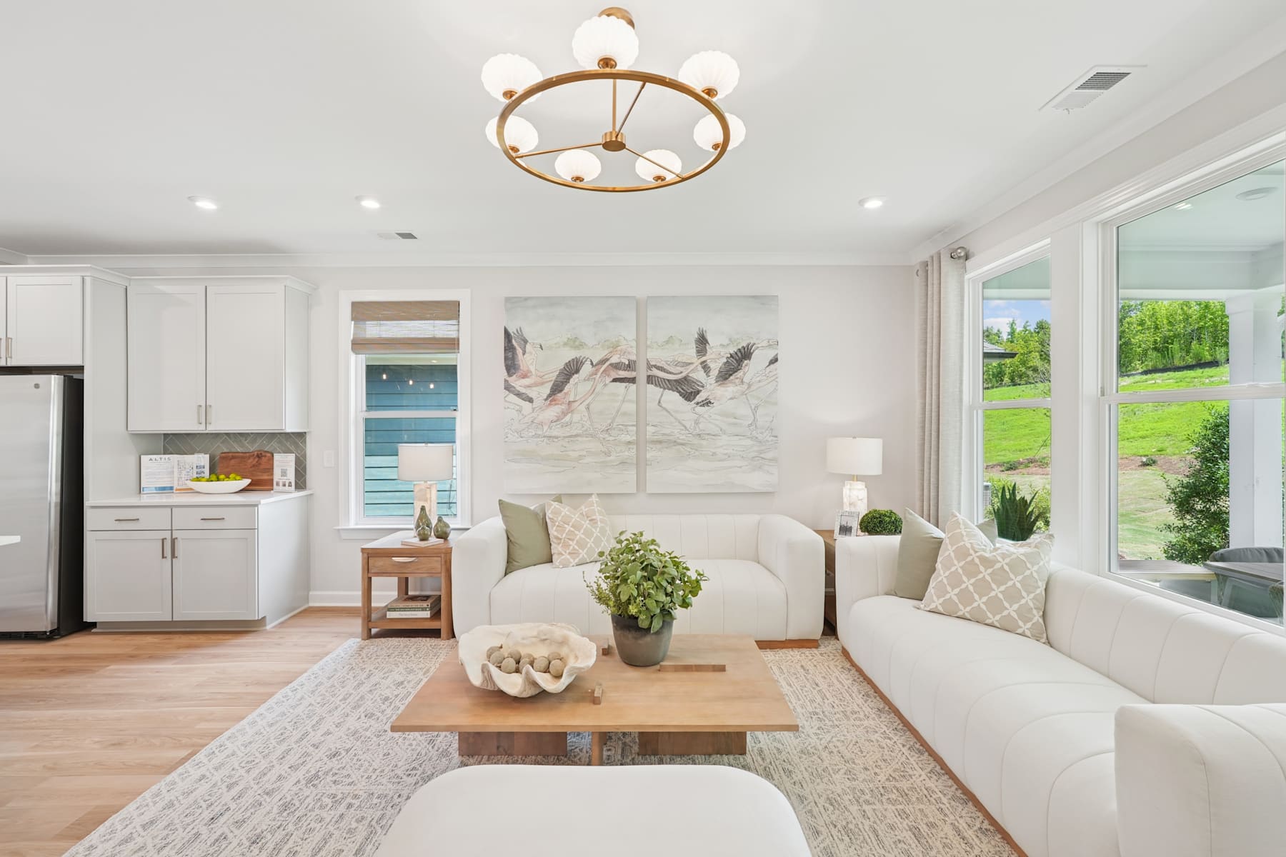 A cozy and bright living room with a large window overlooking a lush outdoor scene, featuring a plush white sofa, a wooden coffee table, and a decorative chandelier overhead.