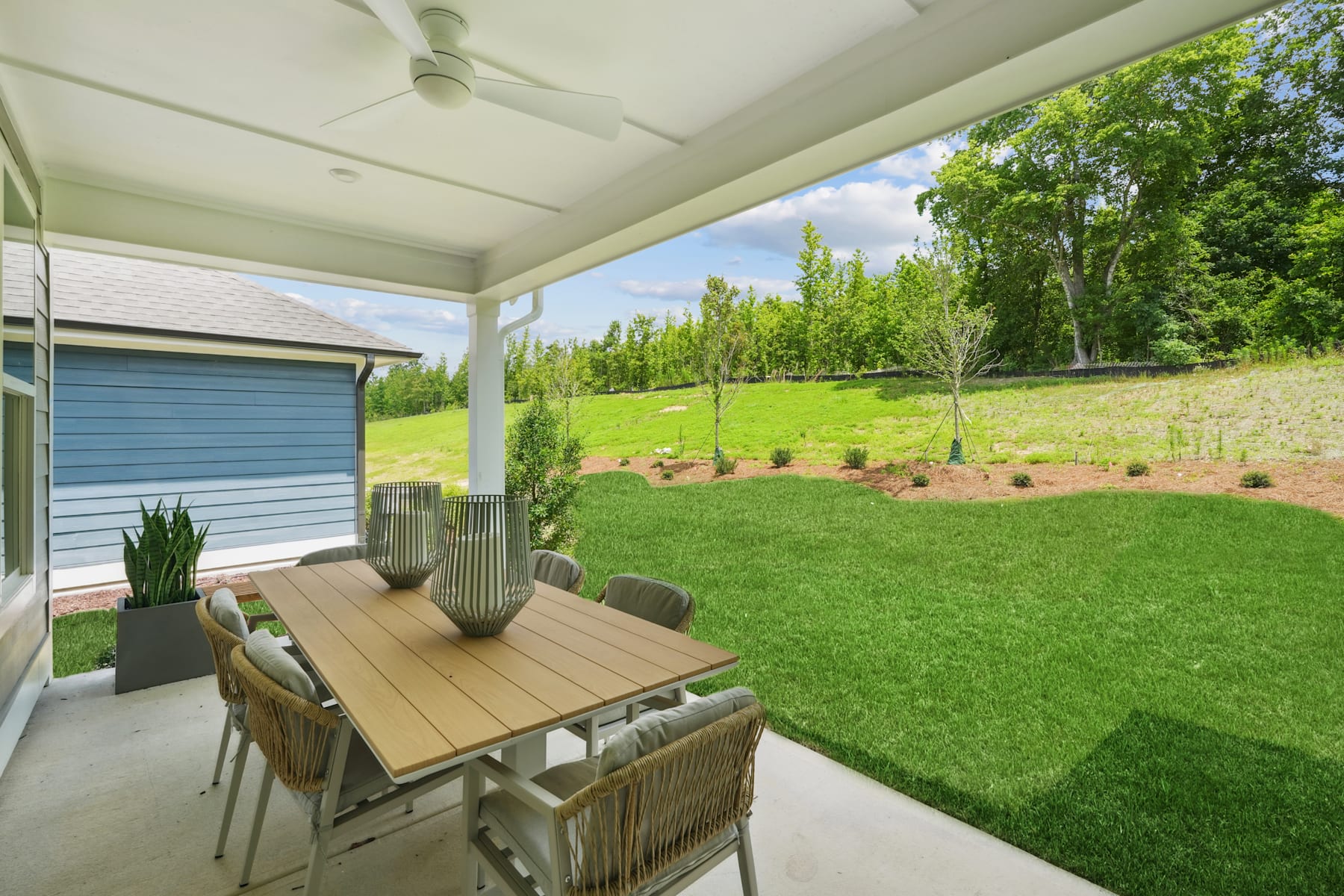 A covered patio with a wooden dining table and chairs overlooking a lush green lawn and trees in the background.