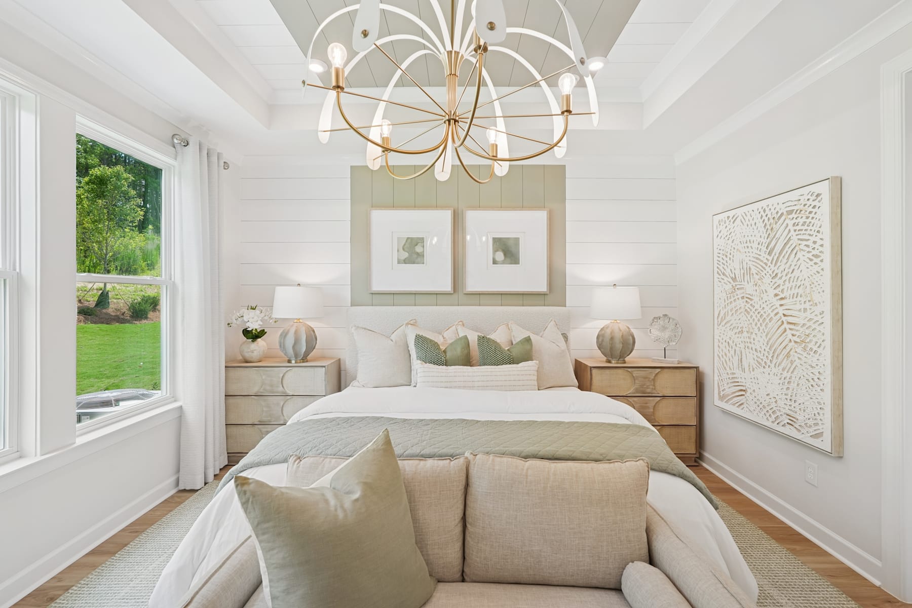 A spacious and elegantly decorated bedroom with a large window overlooking a lush green outdoor landscape, featuring a stylish chandelier, neutral-toned furnishings, and framed artwork on the walls.