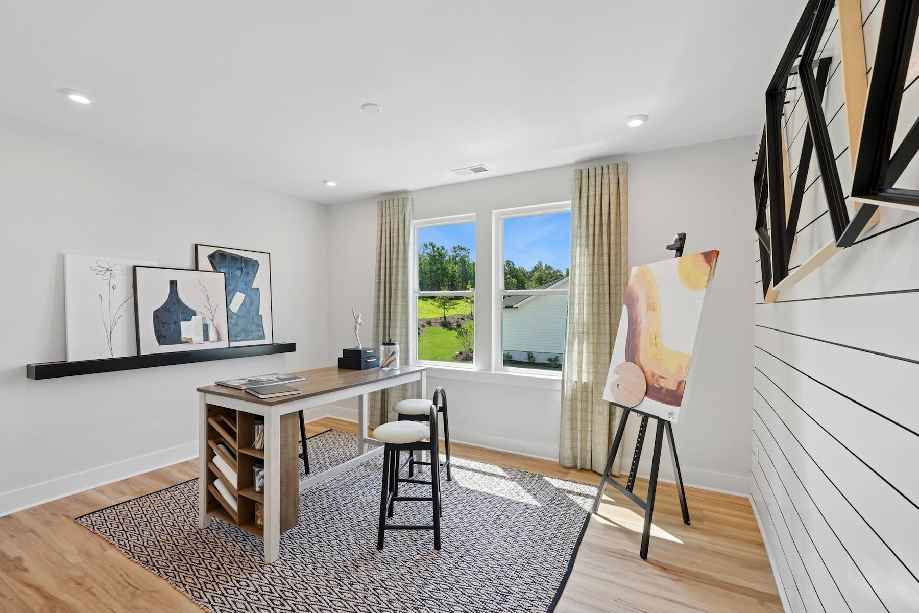 A bright and airy home office space with a wooden desk, artwork on the walls, and a view of the outdoors through a large window.